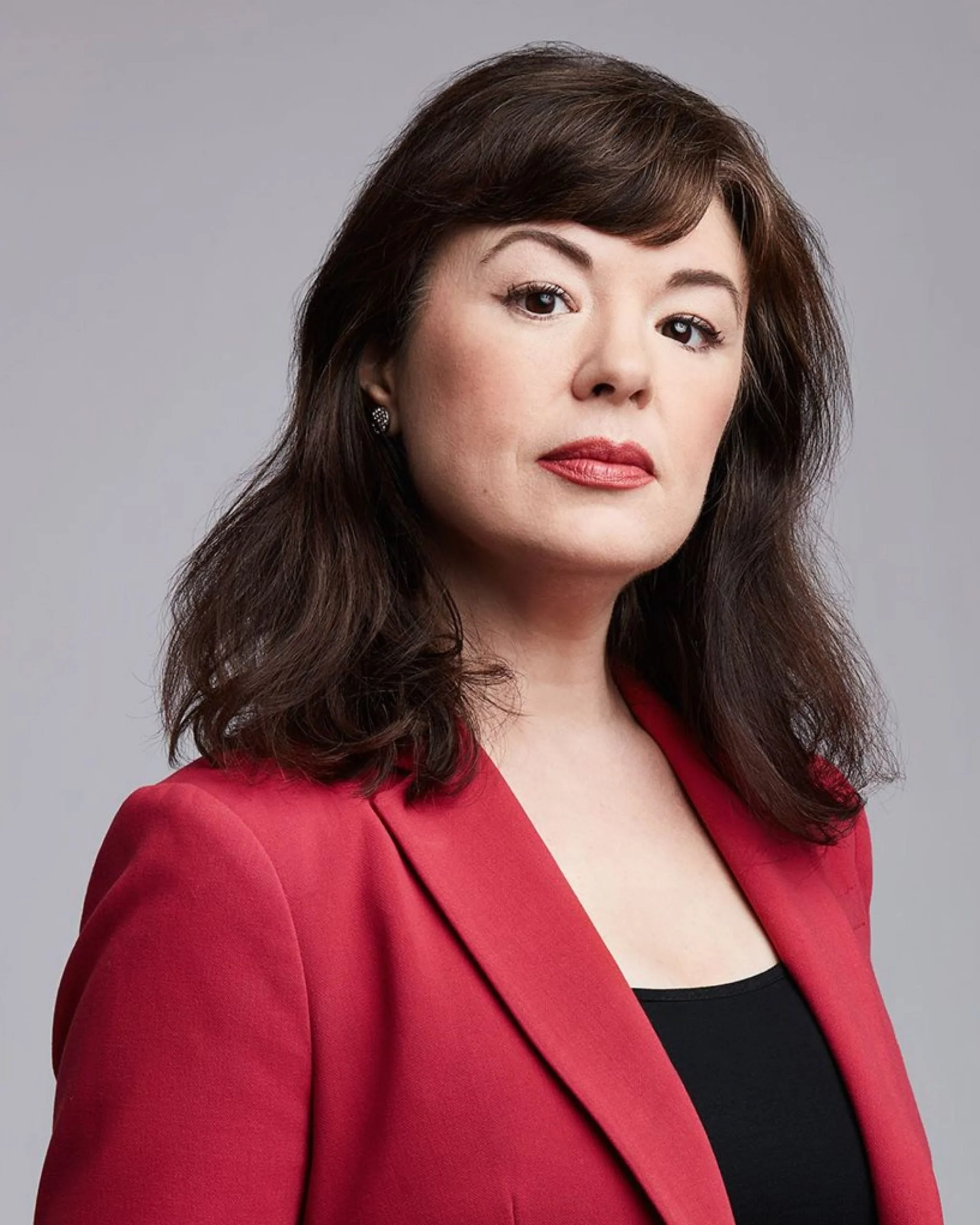 Une femme avec des cheveux bruns ondulés, portant un blazer rouge et un haut noir, regardant sérieusement vers l'avant.