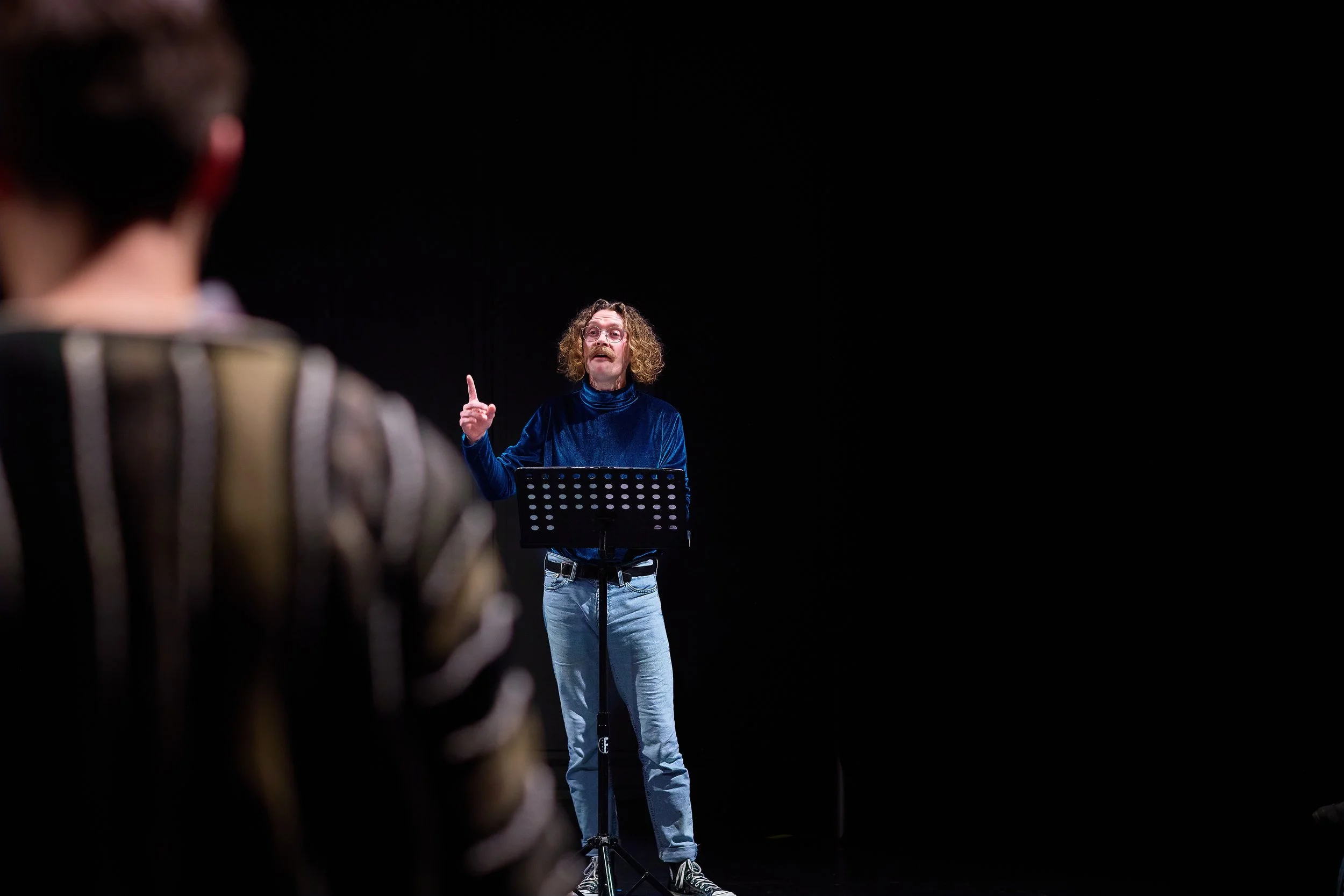 Un homme avec des cheveux bouclés, portant un pull en velours bleu et un jean, se tient devant un micro, faisant une gesture avec un doigt en l'air, dans une pièce sombre, alors qu'une personne en avant de l'image, portant un vêtement à rayures, l'éc