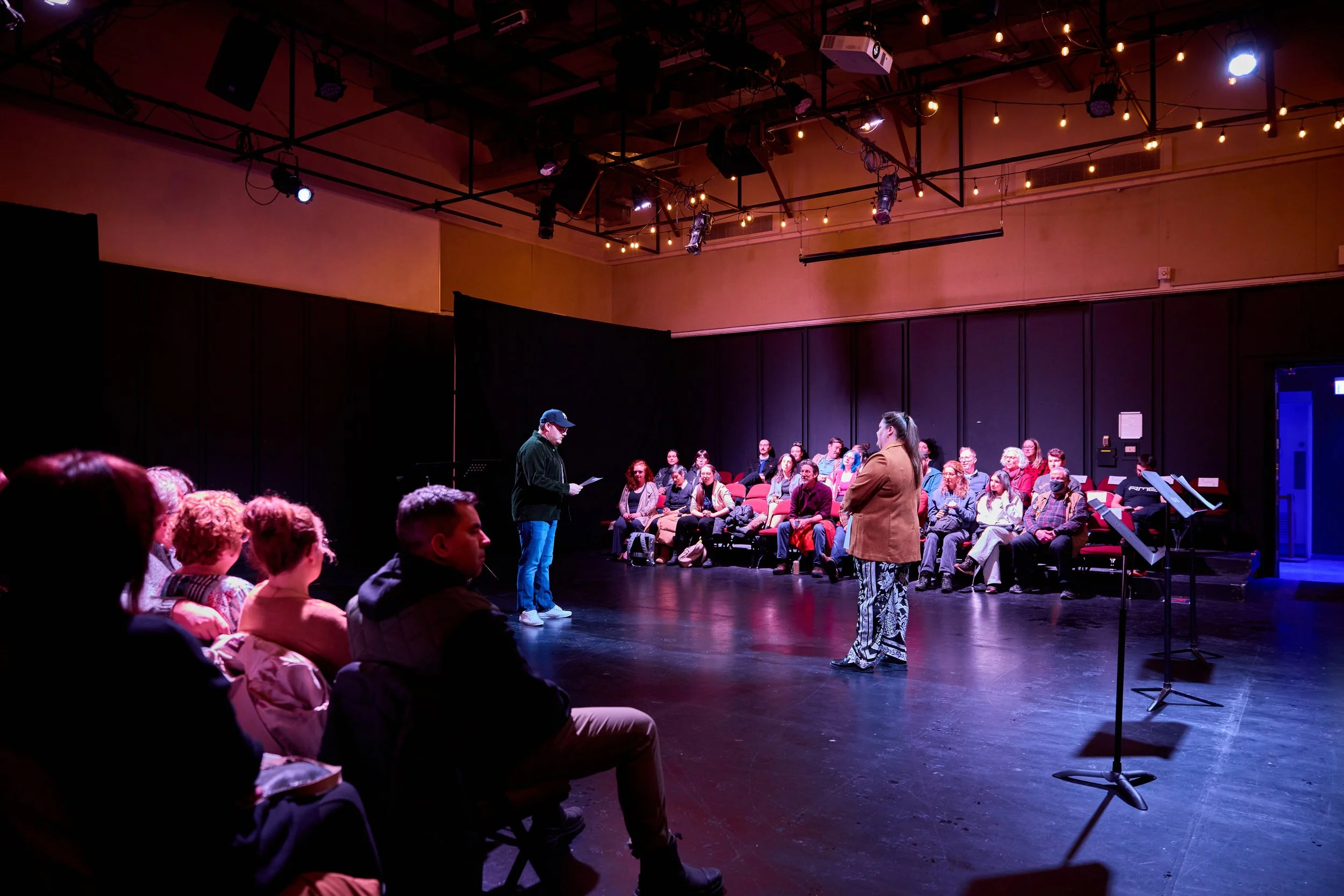 Une pièce de théâtre en cours avec un groupe de spectateurs assis et deux acteurs sur scène, dans une salle sombre avec éclairage suspendu.