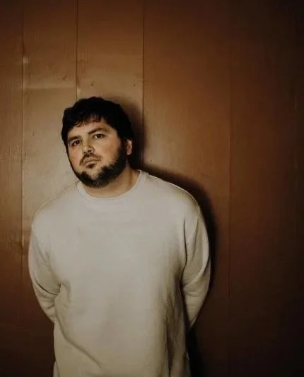Marc-antoine joly. Un homme avec une barbe portant un sweat blanc, debout devant un mur en bois, regardant vers la caméra.