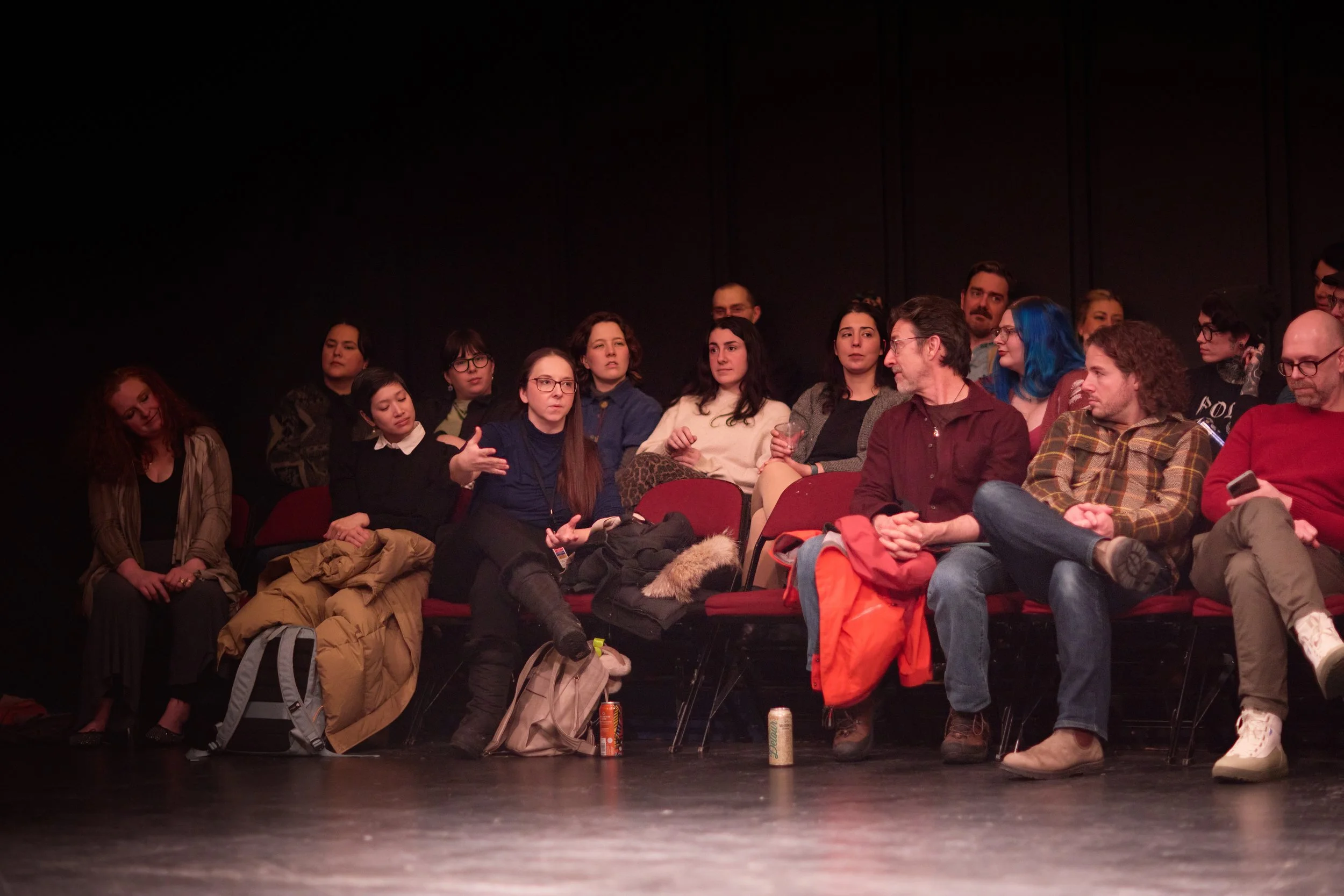 Groupe de personnes assises dans une salle sombre, probablement lors d'un événement ou d'une discussion, avec des bagages et des boissons devant elles.