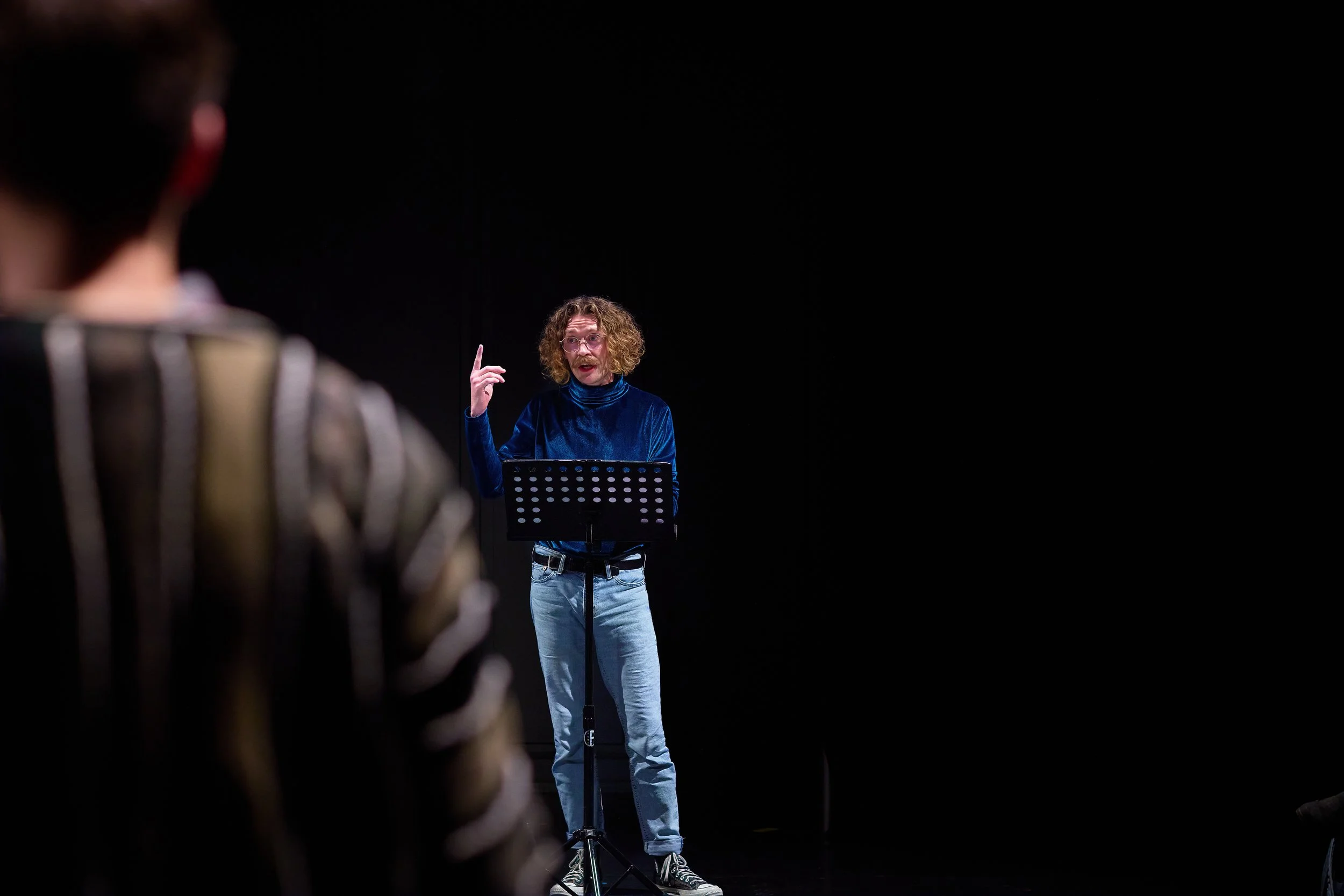 Un homme avec des cheveux bouclés et roux, portant un chandail en velours bleu et un jean clair, parle en direction d'un groupe, avec un support de paroles devant lui, dans une pièce sombre.