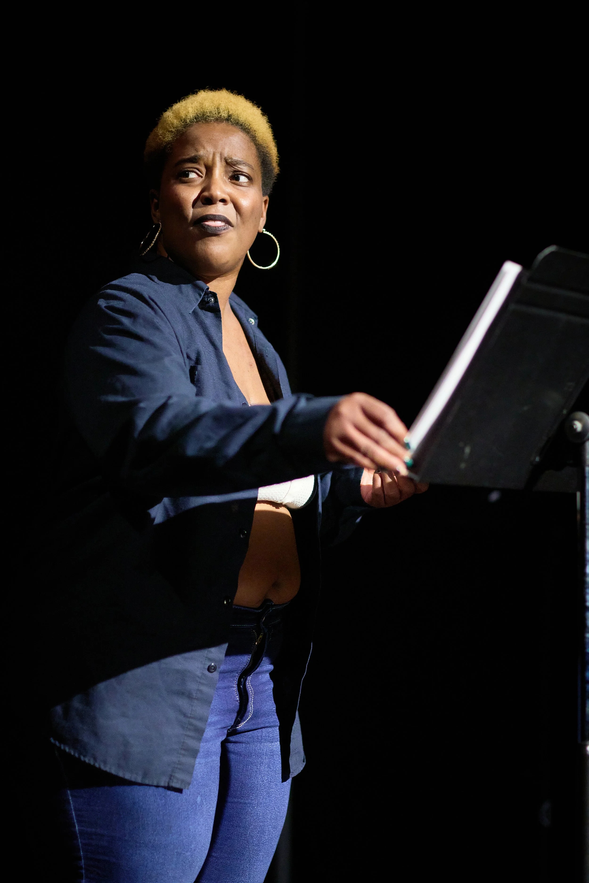 Une femme avec des cheveux blonds courts et un visage expressif regarde vers la gauche, tenant un carnet ou un livre. Elle porte un chemisier bleu foncé et des boucles d'oreilles en cercle. L'arrière-plan est noir.