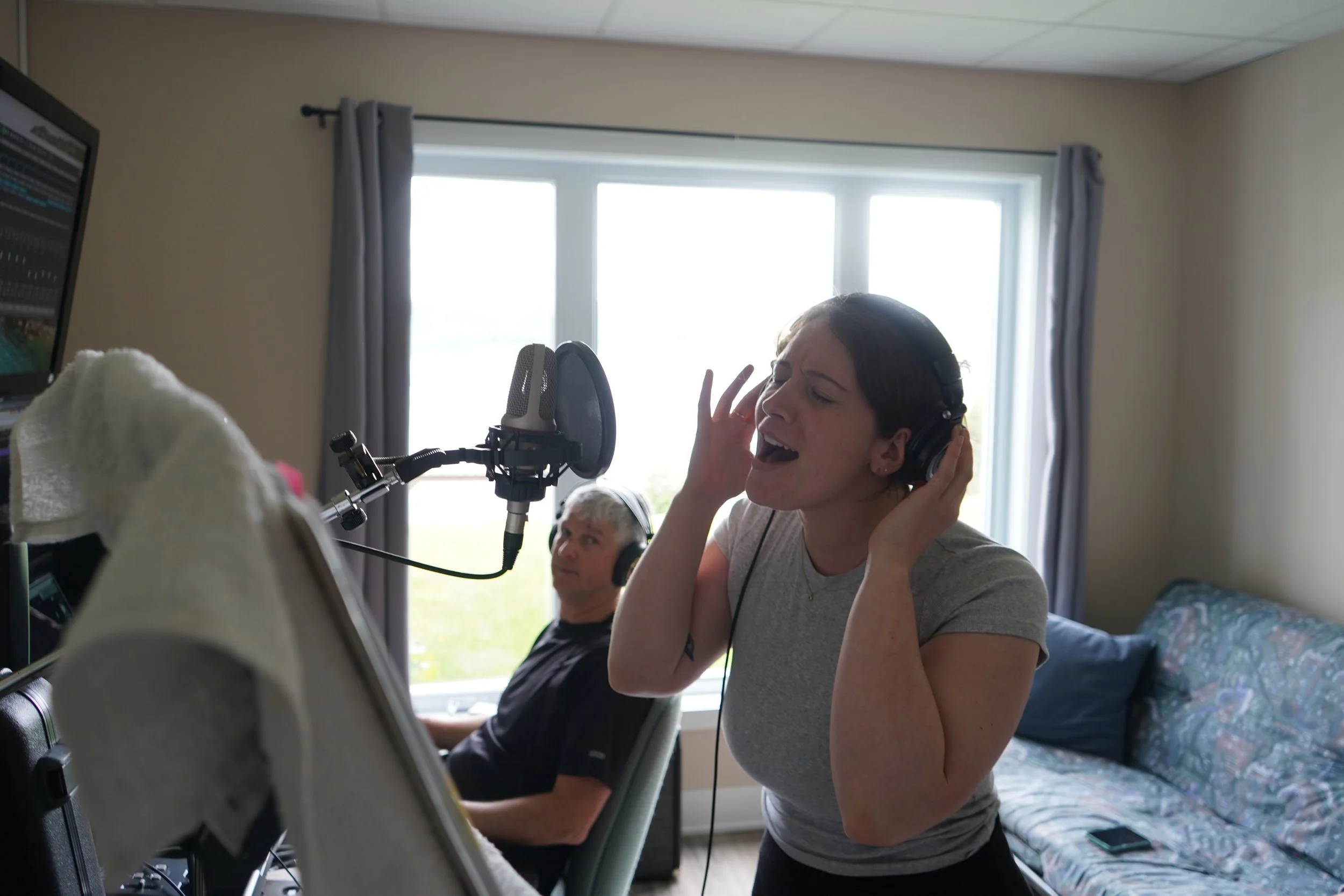 Une jeune femme chante et enregistre dans un studio à la maison, avec un homme assis en arrière-plan. La pièce est éclairée par la lumière naturelle d'une fenêtre derrière eux.