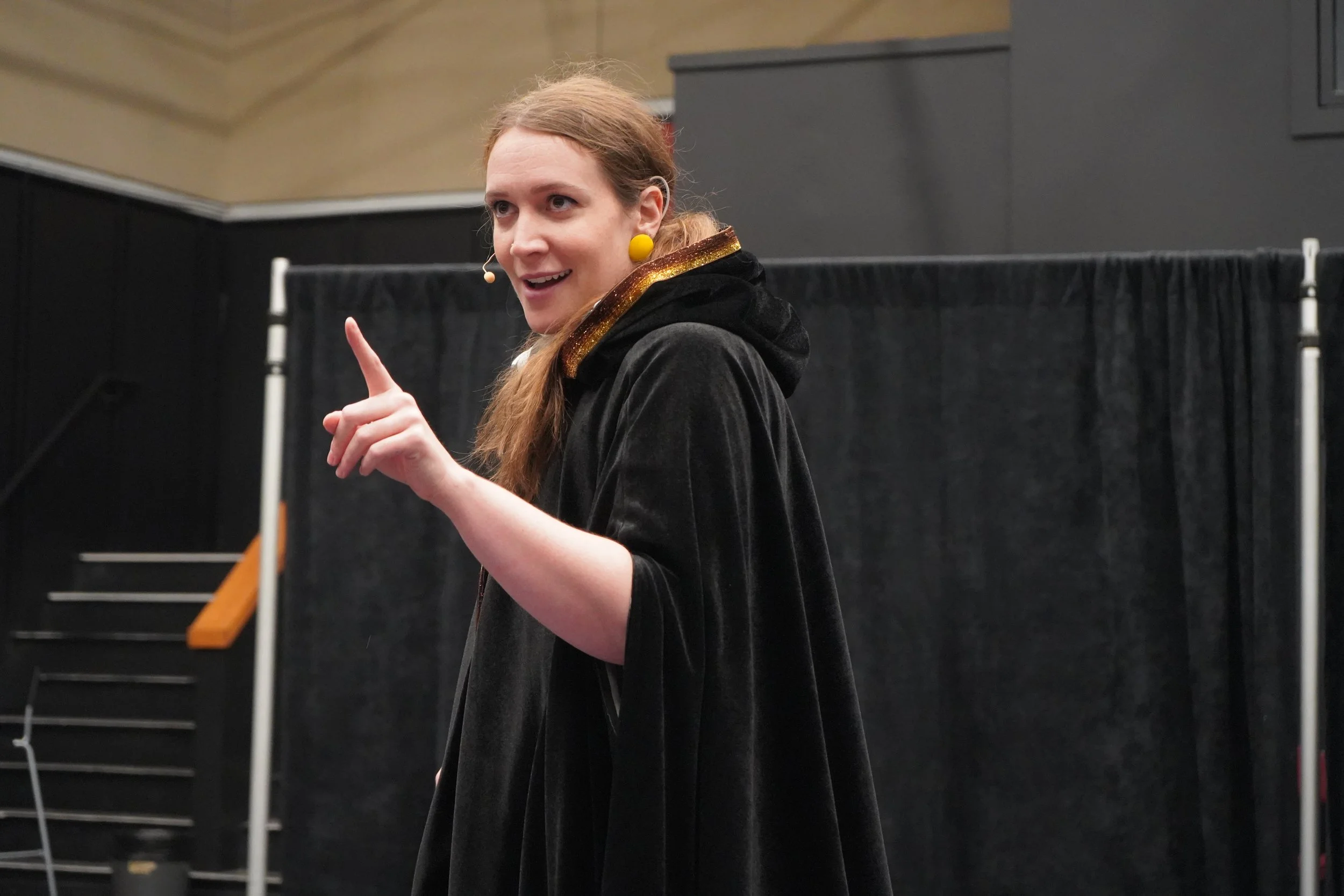 Une femme avec des cheveux bruns, portant une robe ou une cape noire avec des accents dorés, parlant ou faisant un geste de la main lors d'une présentation ou d'une performance sur scène.