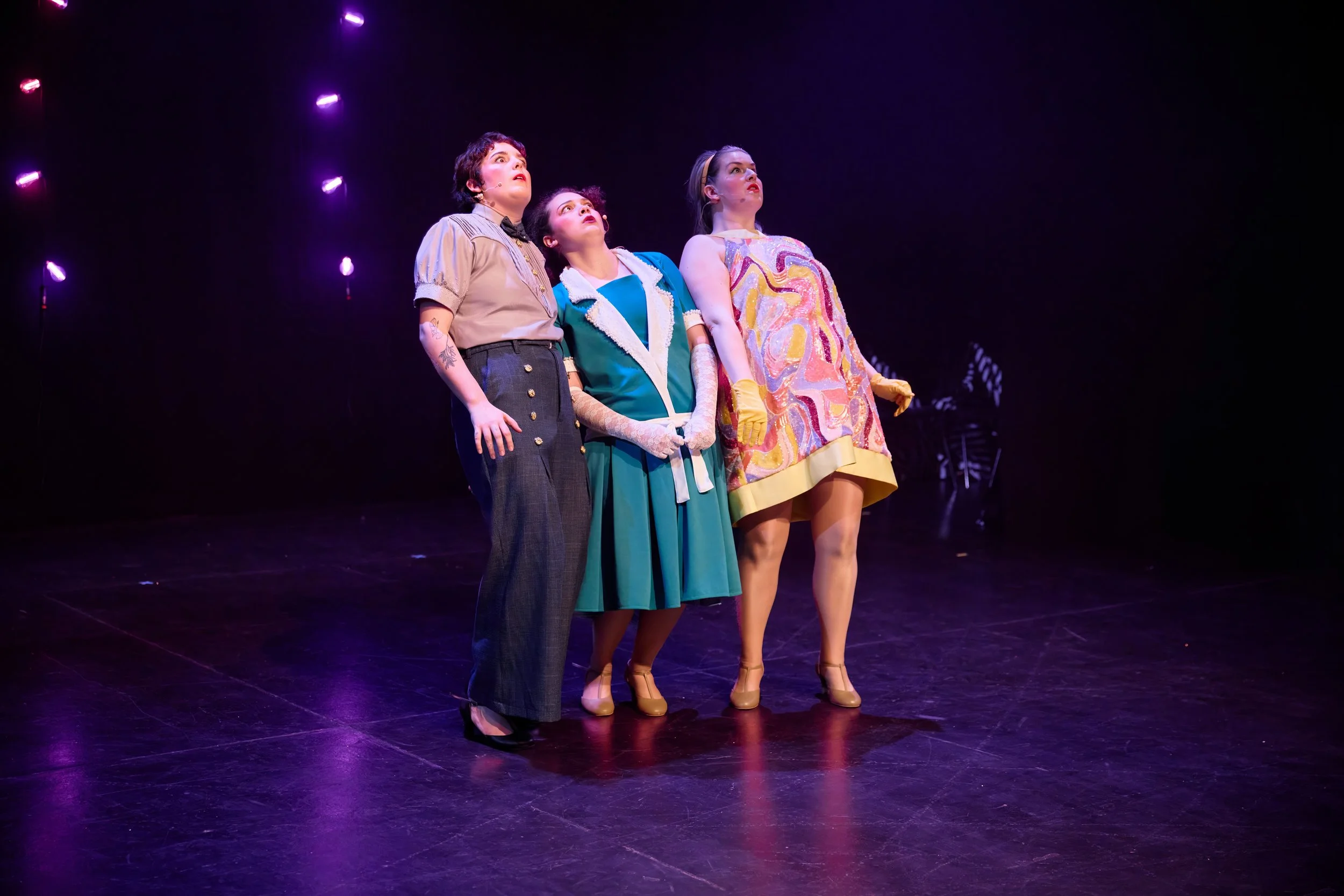 Trois femmes en costumes vintage sur scène sous des lumières violettes, regardant vers le haut.