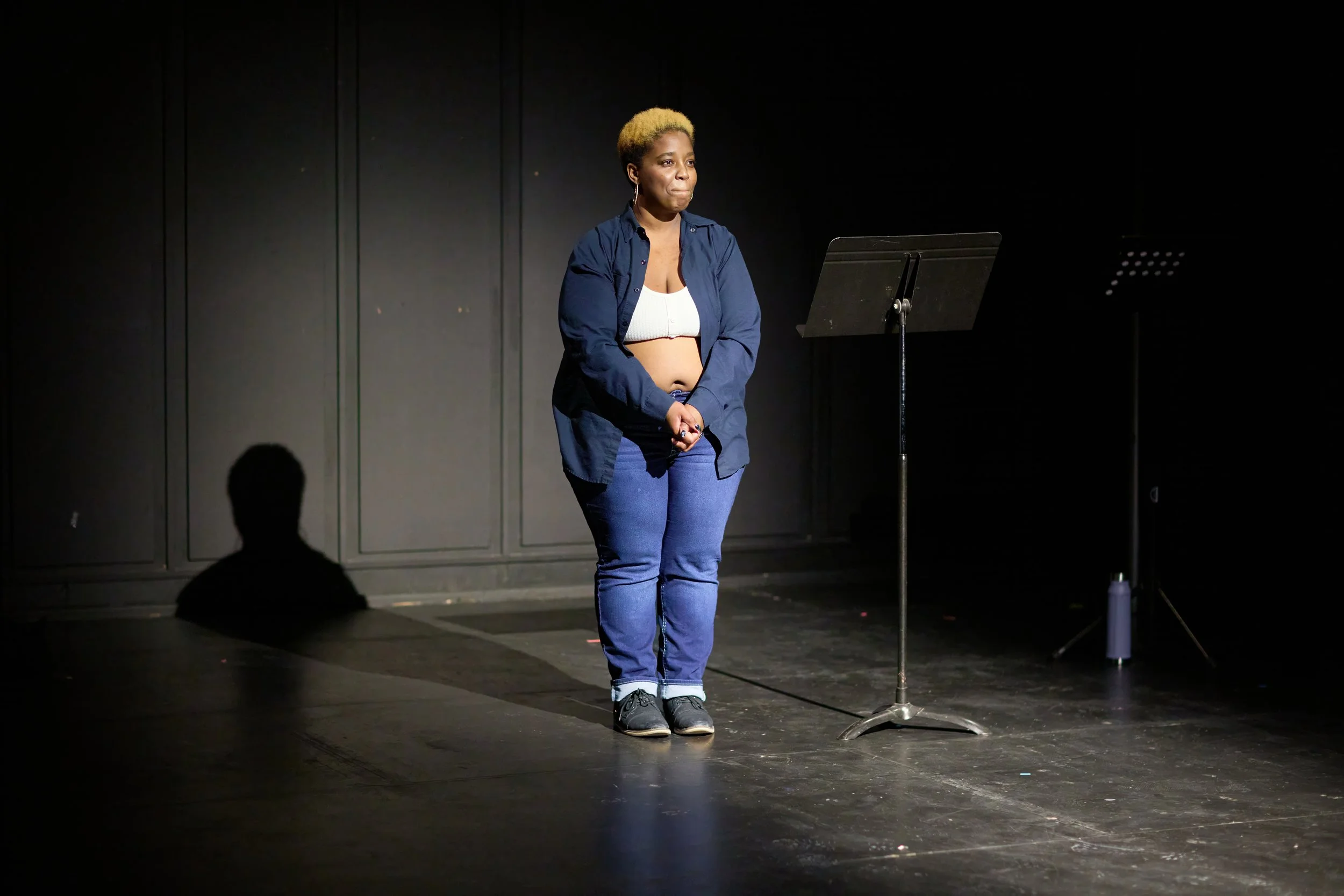 Une femme debout sur une scène sombre, portant un haut blanc, une chemise bleue ouverte, et un pantalon bleu, avec un pupitre devant elle.