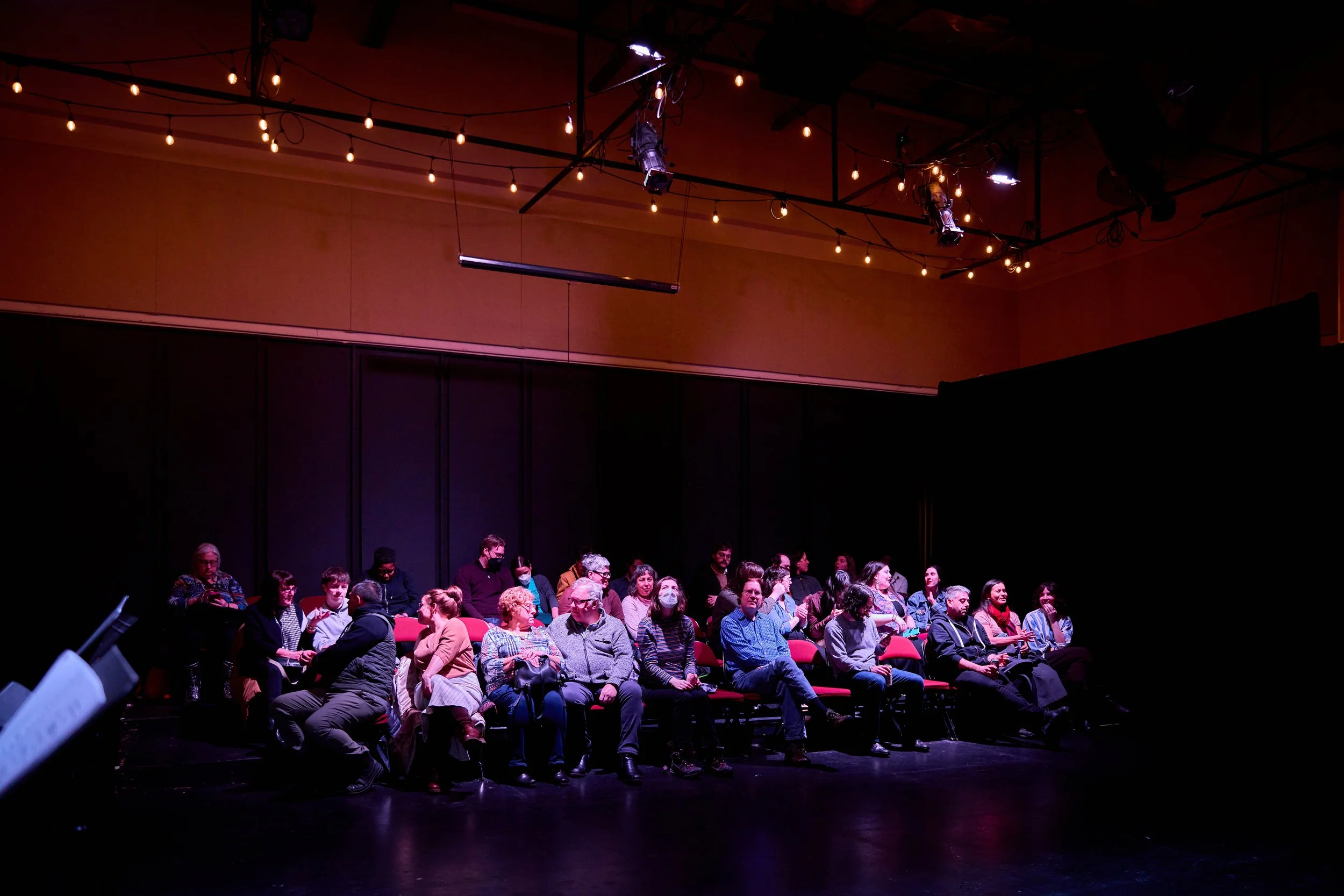 Groupe de personnes assises dans une salle sombre avec un éclairage tamisé, portant des masques faciaux, probablement un théâtre ou un auditorium.