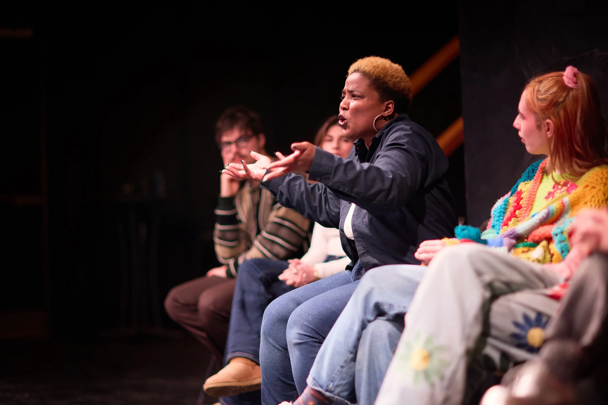 Une femme parle passionnément lors d'une discussion ou d'une performance, entourée de plusieurs personnes qui écoutent attentivement.