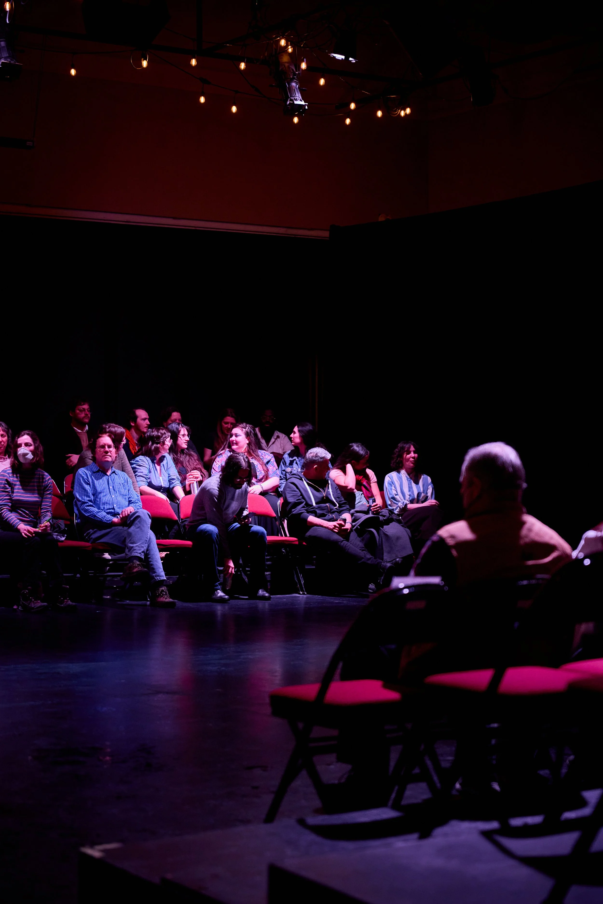 Groupe de personnes assises dans une salle de théâtre ou salle de spectacle, attentives à une performance ou une présentation, sous un éclairage tamisé avec des ampoules suspendues au plafond.