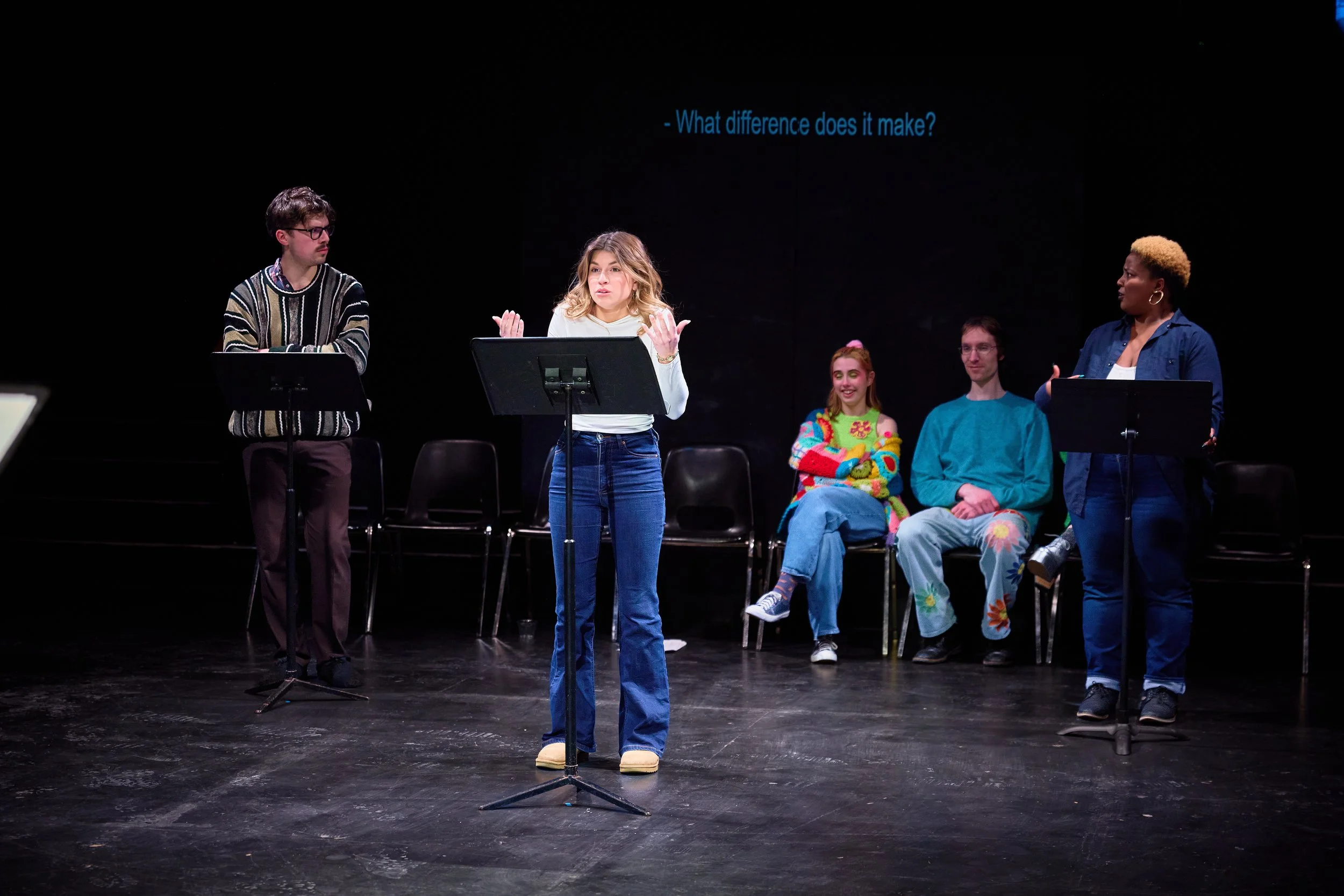 Groupe de cinq personnes en train de jouer une pièce de théâtre ou de faire un atelier sur scène, avec écrans en arrière-plan affichant le texte 'What difference does it make?'.