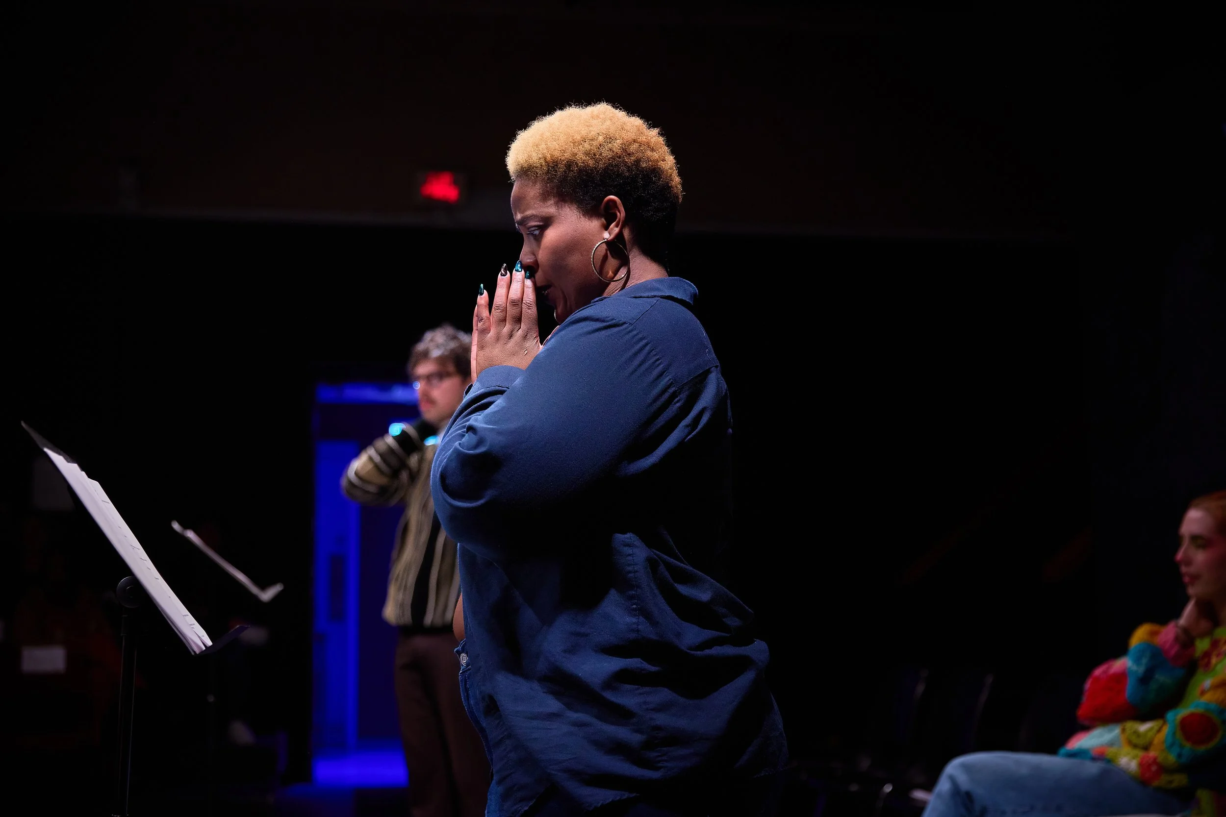 Une femme avec des cheveux courts éclaircis prie dans une pièce sombre, d'autres personnes sont présentes en arrière-plan, dont un homme en train de parler et une femme assise à droite en vêtement coloré.