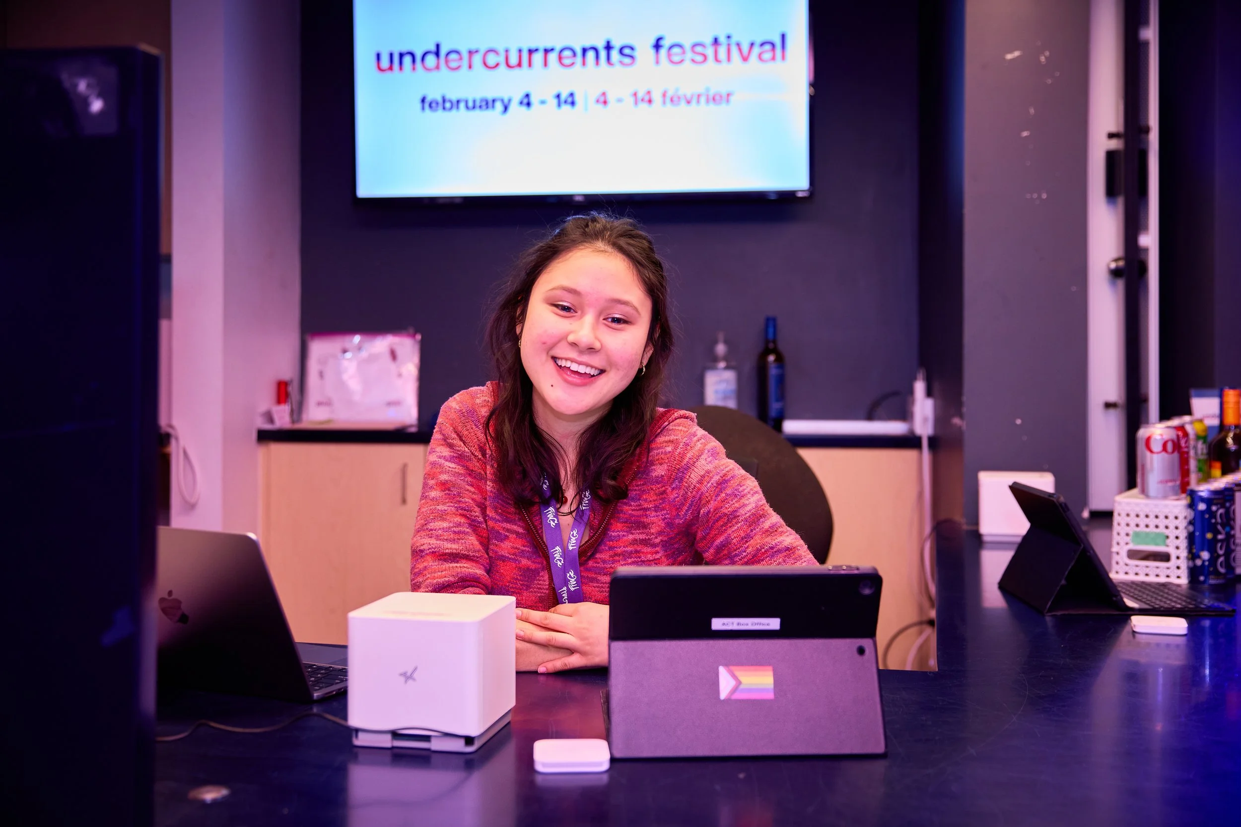 Jeune femme souriante devant un ordinateur lors du festival Undercurrents, affiché sur un écran derrière elle, avec boissons et appareils électroniques sur la table.