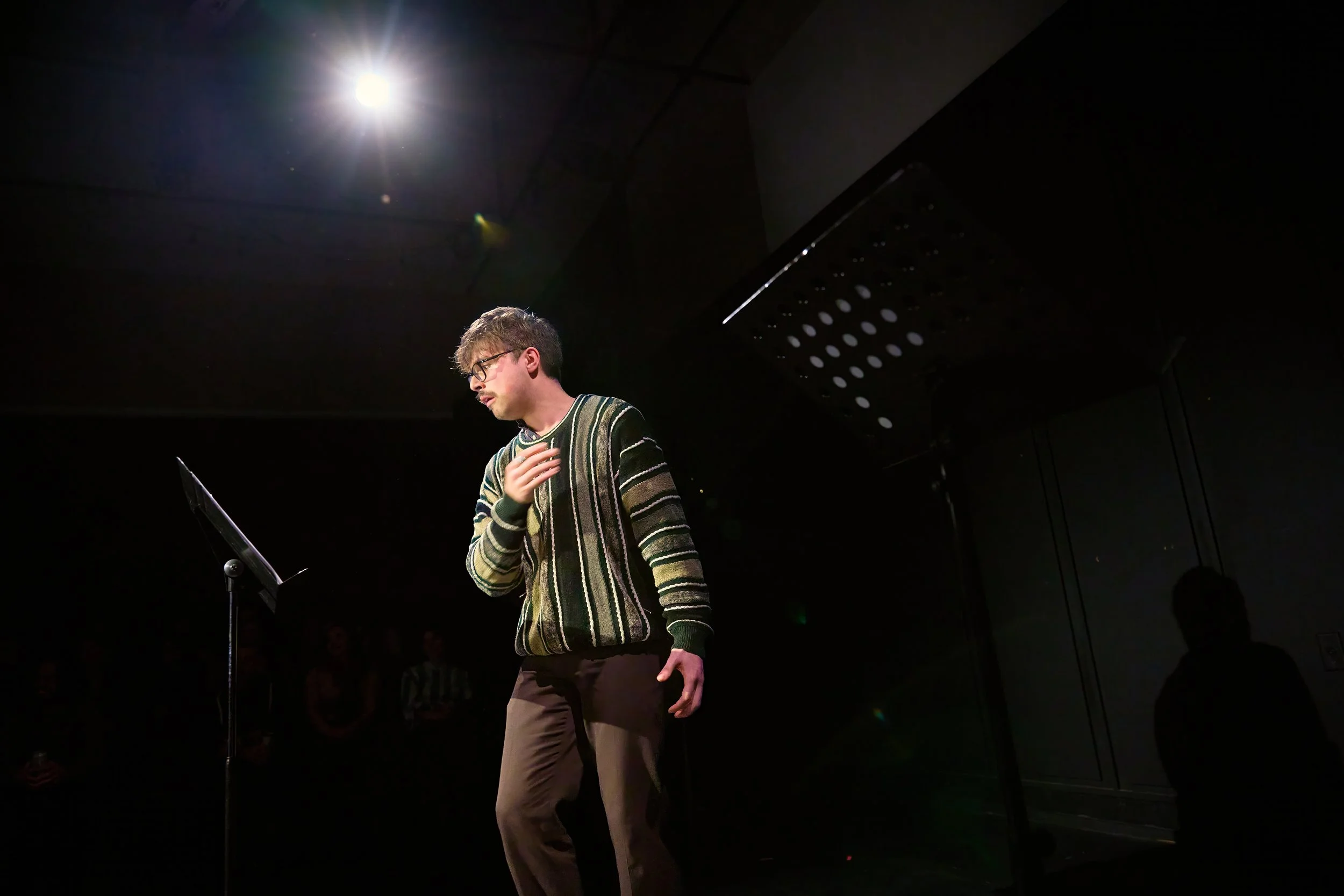 Un jeune homme portant un pull rayé, debout devant un pupitre avec un microphone, sous un éclairage de scène, en train de lire ou de réciter.