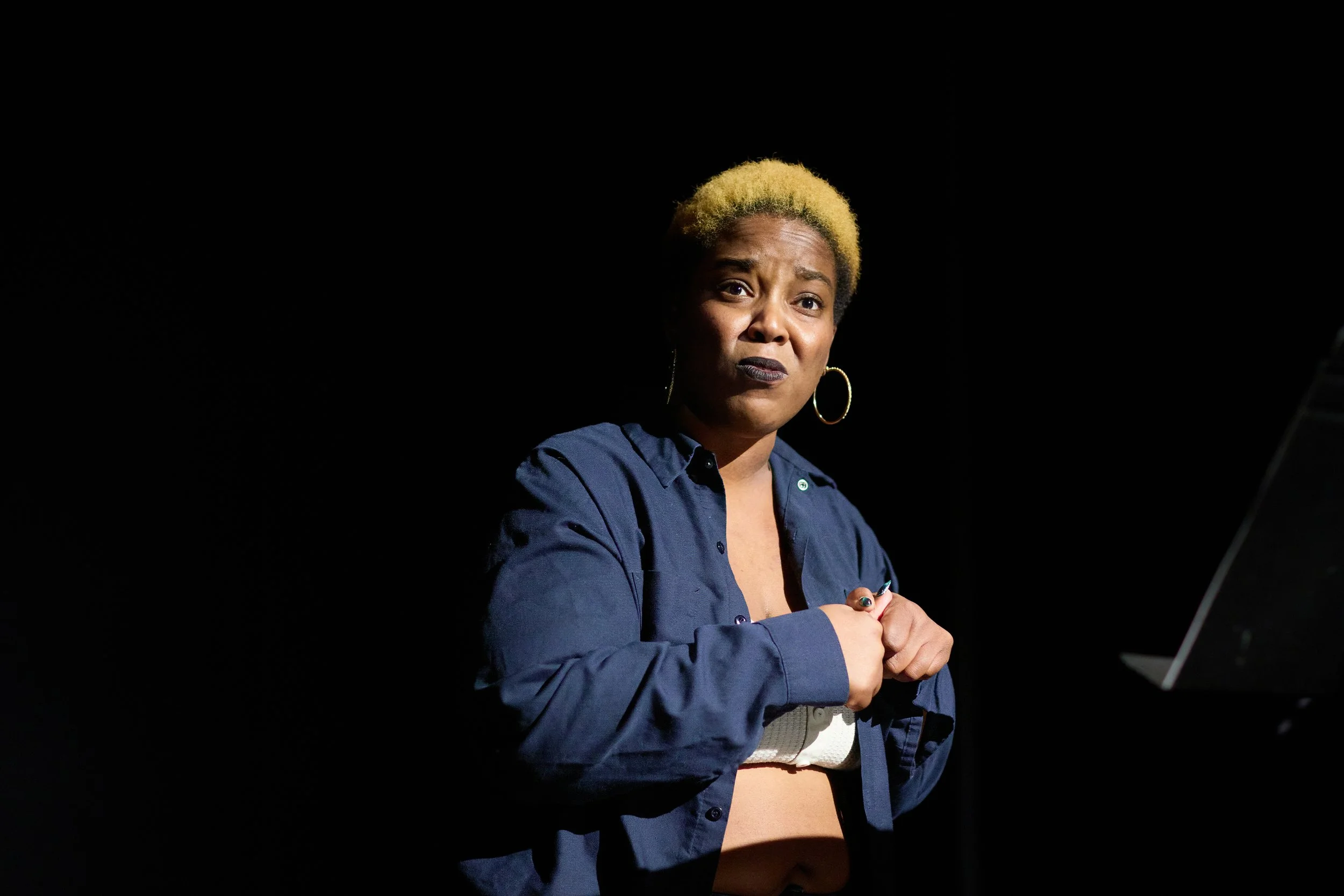 Une femme avec des cheveux blonds courts portant un jean bleu et une pièce de vêtement blanche, debout dans un fond noir, regardant la caméra avec une expression sérieuse.