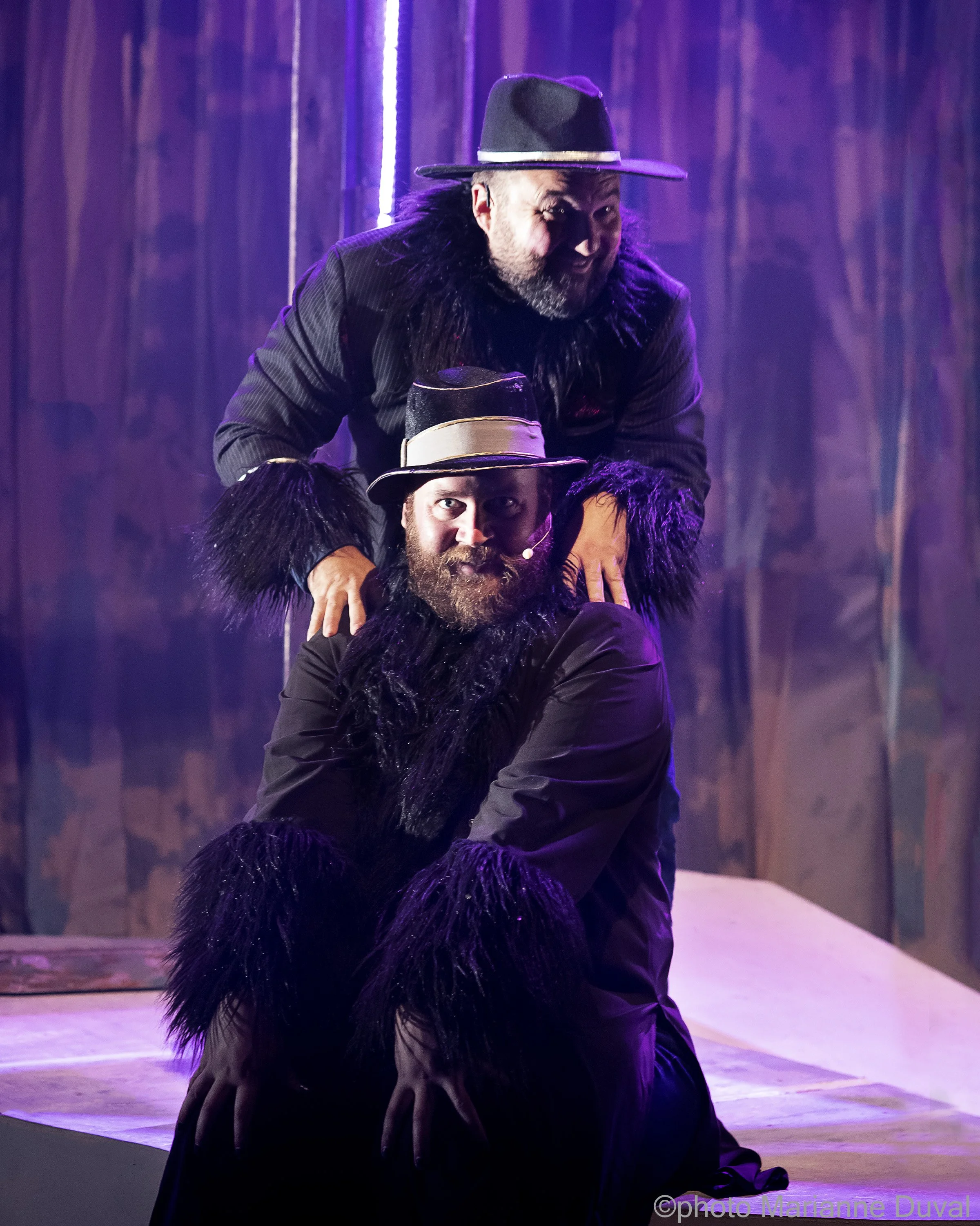 Deux hommes avec des costumes noirs, des chapeaux et des détails en plumes noires posent de manière théâtrale sur scène avec un arrière-plan en bois et des lumières violettes.