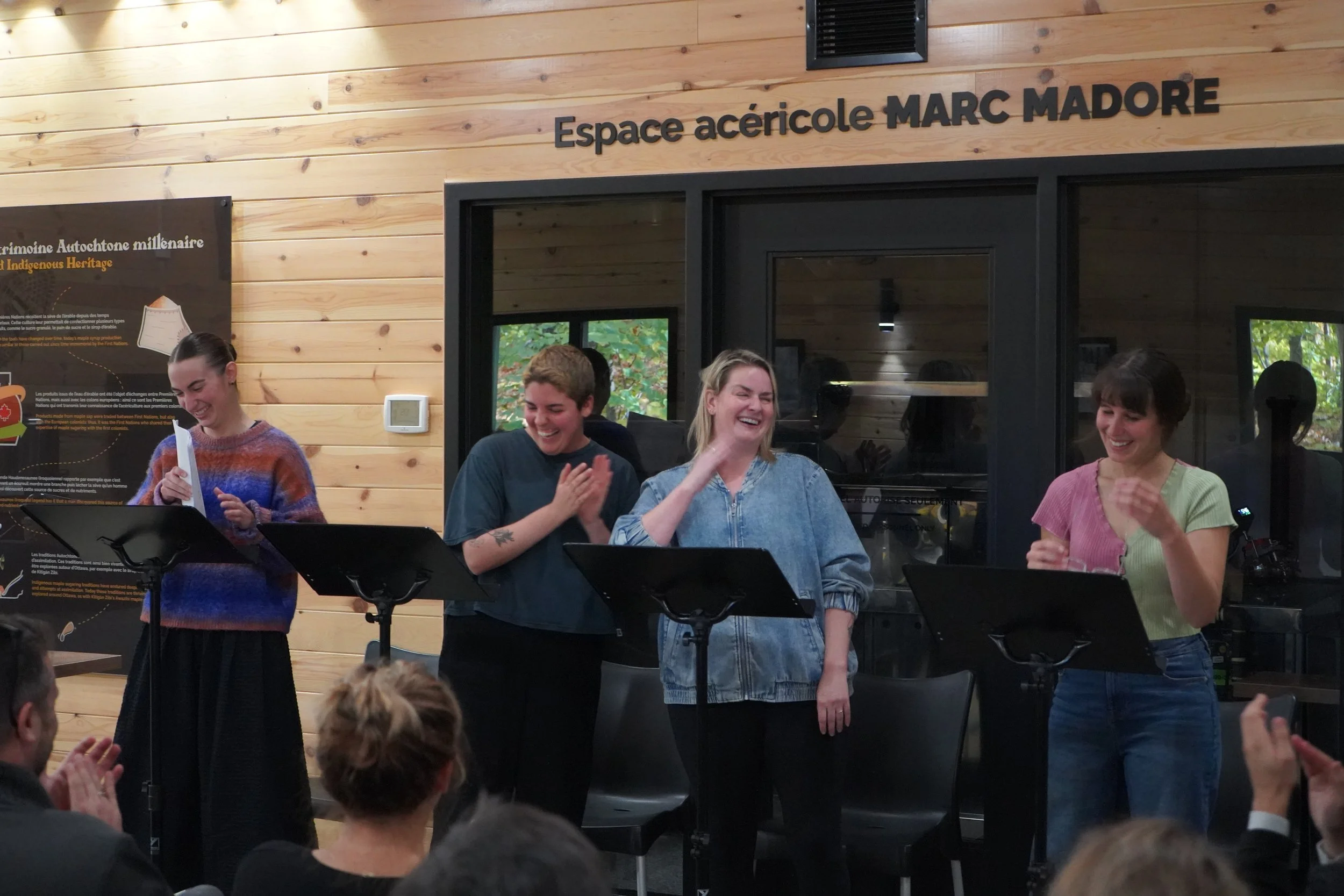 Cinq femmes souriantes et riant lors d'une performance ou d'une présentation, debout derrière des pupitres musicaux dans une salle avec un mur en bois et la signalisation "Espace acériole Marc Madore".