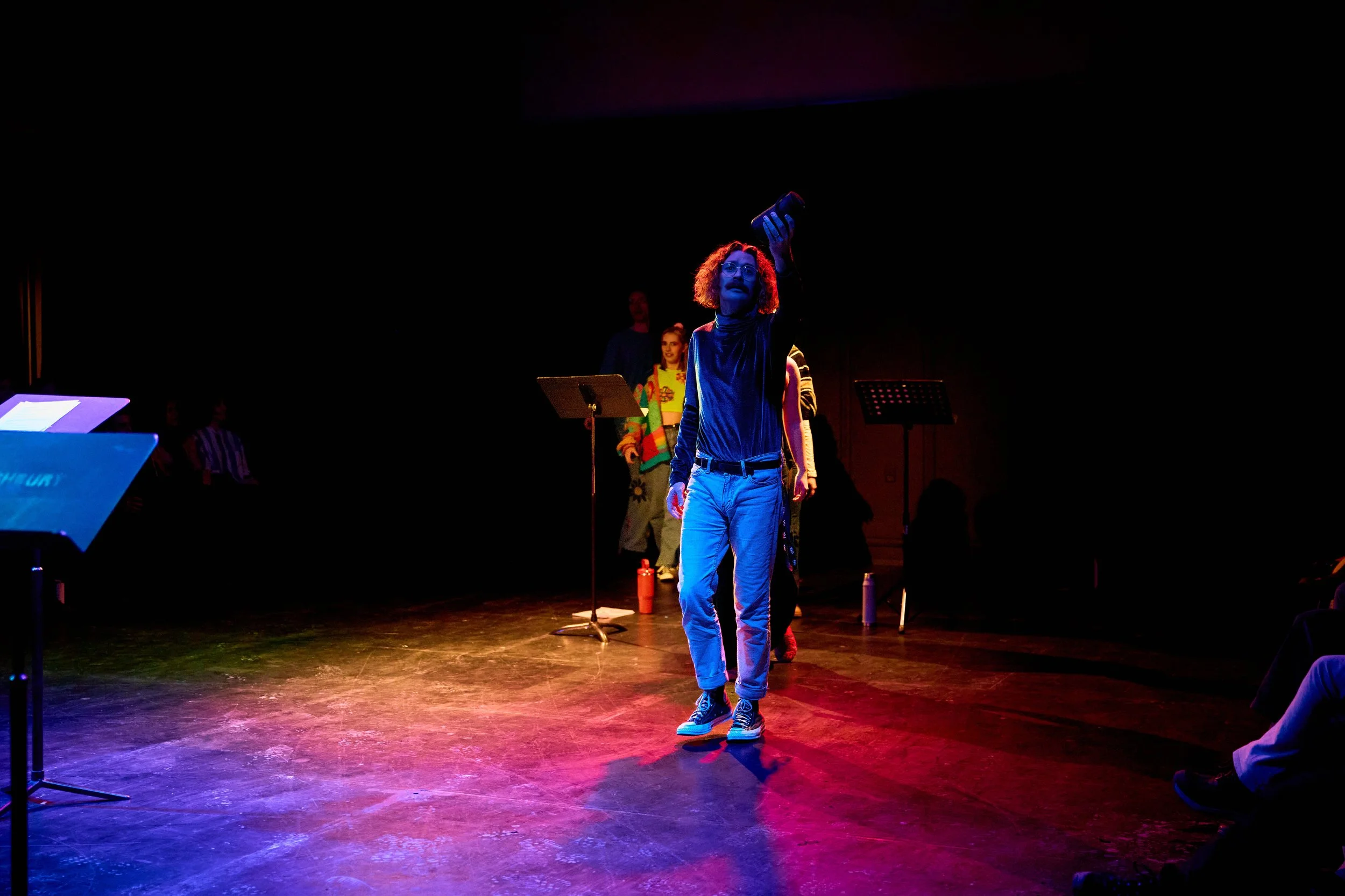 Personne sur scène, tenant un téléphone, avec d'autres personnes en arrière-plan, éclairage coloré