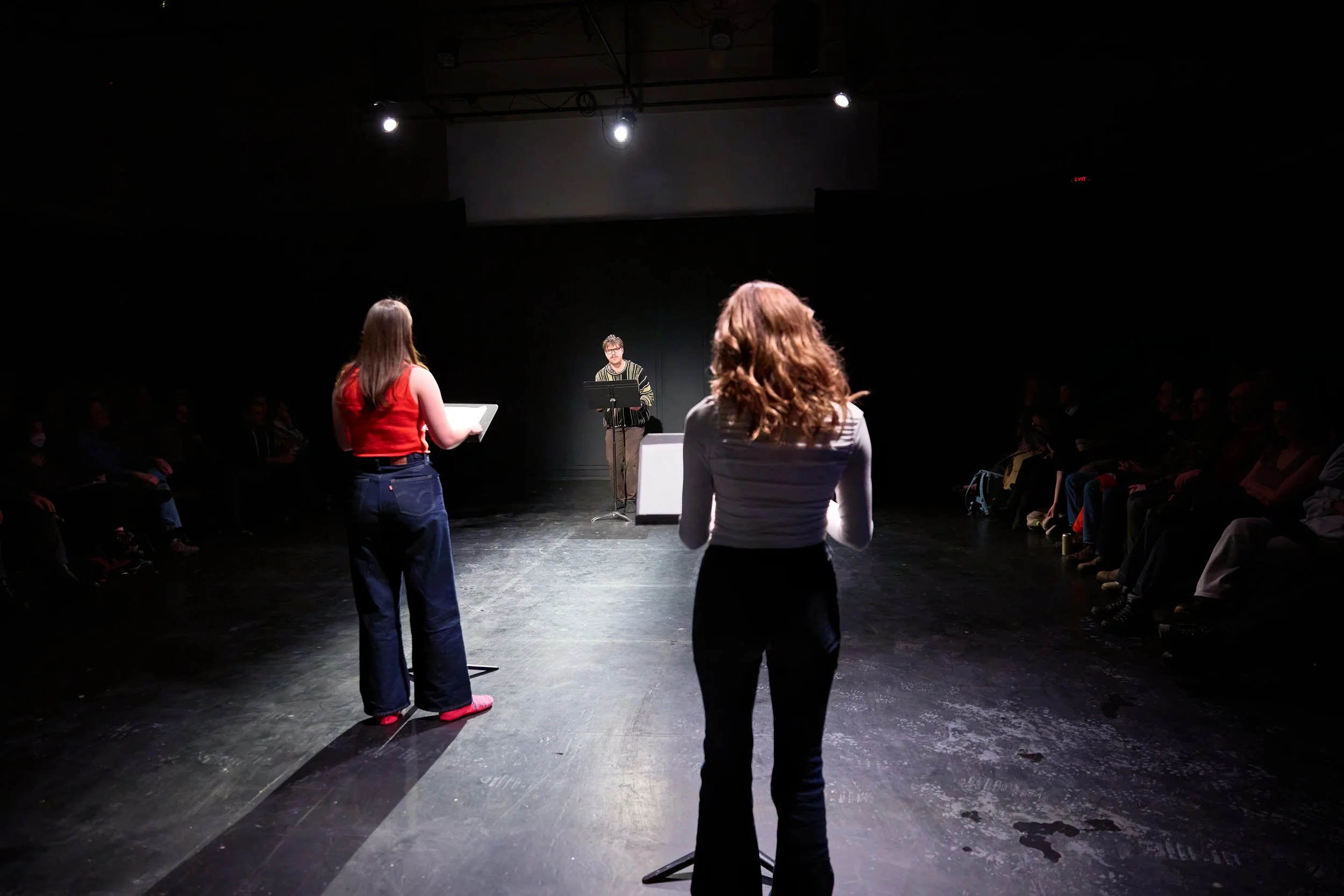 Une scène de théâtre ou de lecture avec deux femmes au premier plan, une lisant sur un pupitre et l'autre en face, un homme au centre derrière un pupitre, un public assis dans l'obscurité.