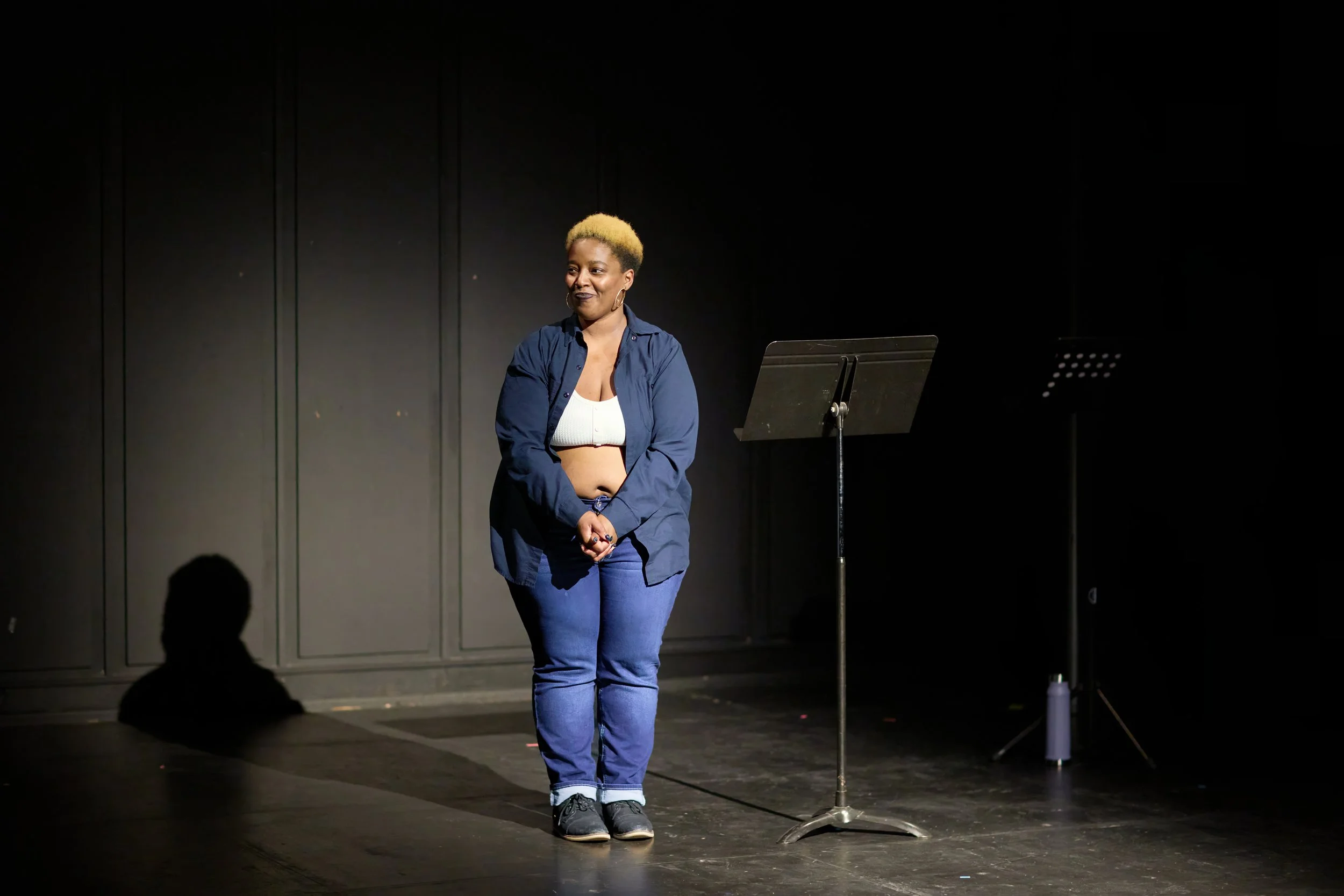 Une femme debout sur scène sous un éclairage sombre, porte un jean et une veste bleue, à côté d'un pupitre, dans une atmosphère de spectacle ou de lecture