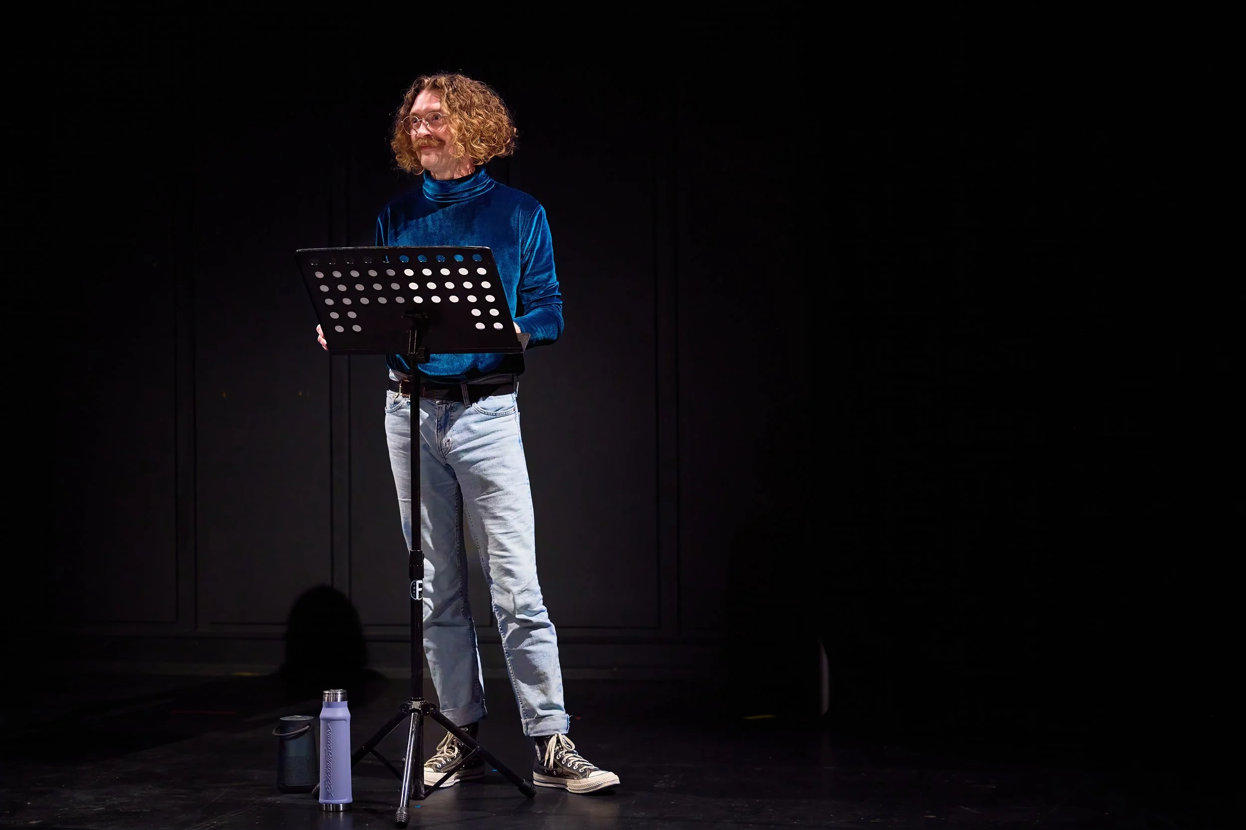 Un homme avec des cheveux bouclés, porter un sweat bleu, debout à un micro avec une partition, éclairé sur un fond noir.