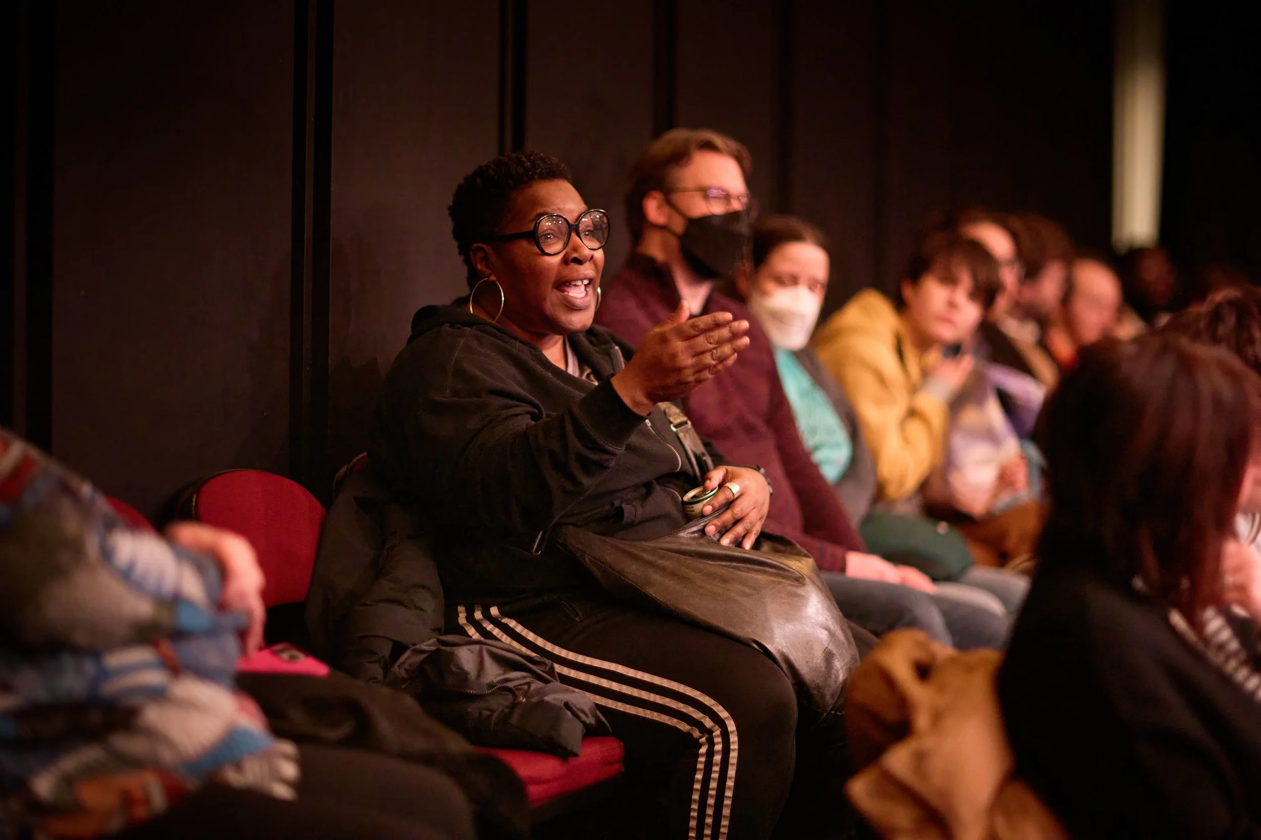 Une femme noire portant des lunettes et une veste noire parle en levant la main lors d'une réunion ou conférence, entourée de personnes portant des masques faciaux assises sur des sièges rouges.