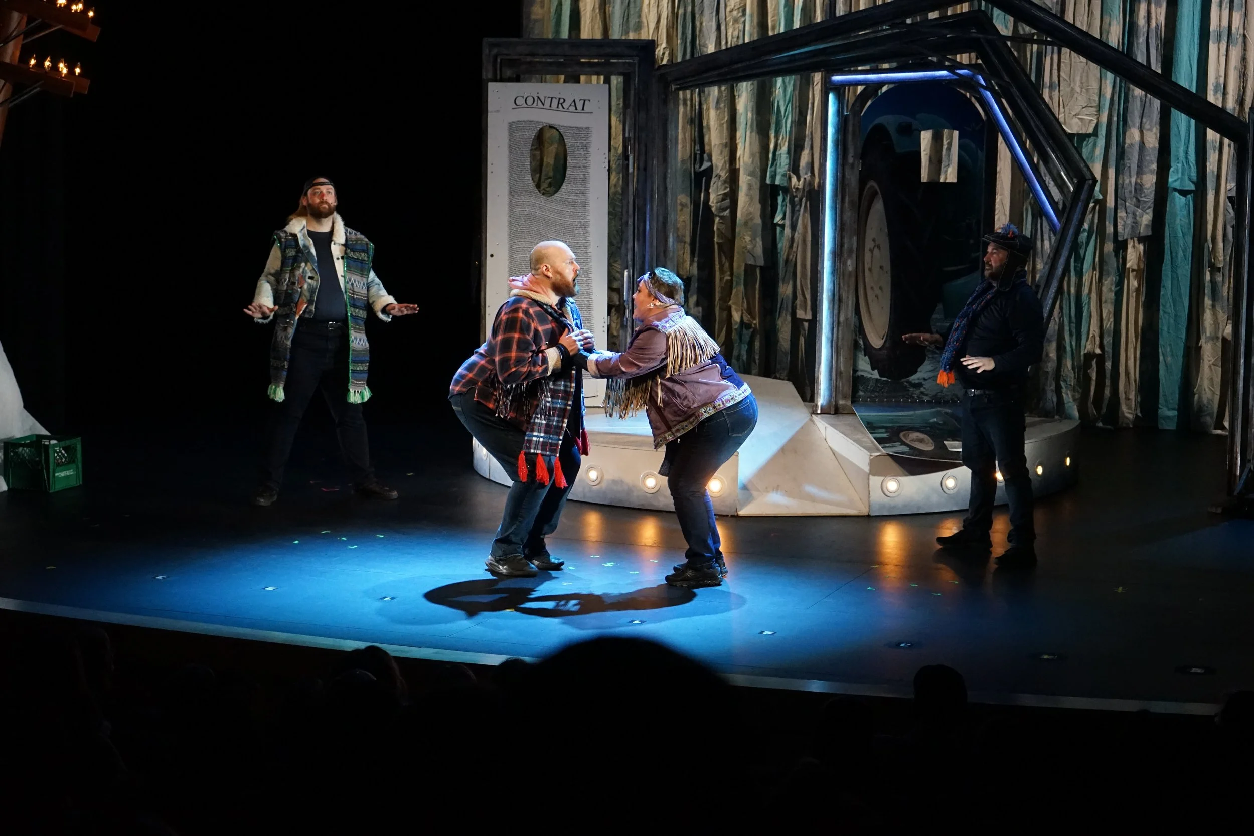 Scène de théâtre avec quatre acteurs, deux au centre en costume décontracté, deux autres en costume plus sombre, sur un décor évoquant une scène de jeu ou de spectacle.