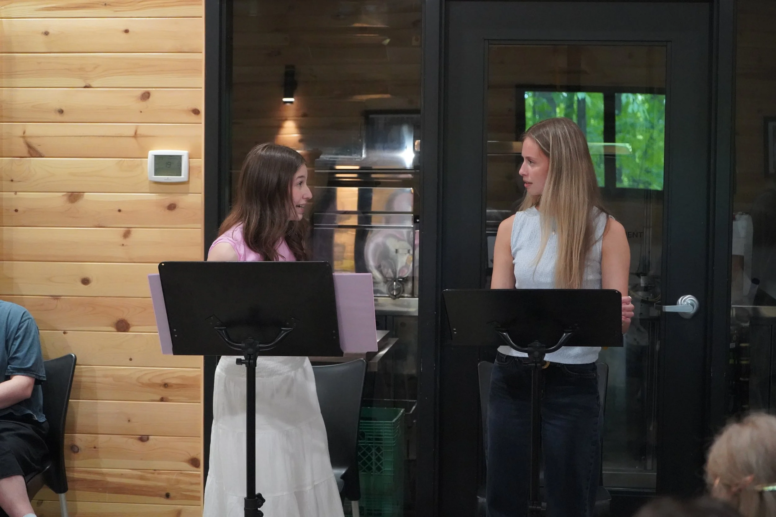 Deux femmes discutent dans une salle intérieure avec des murs en bois et une grande porte en verre. Il y a un mur avec un thermostat et des partitions pour musiciens.