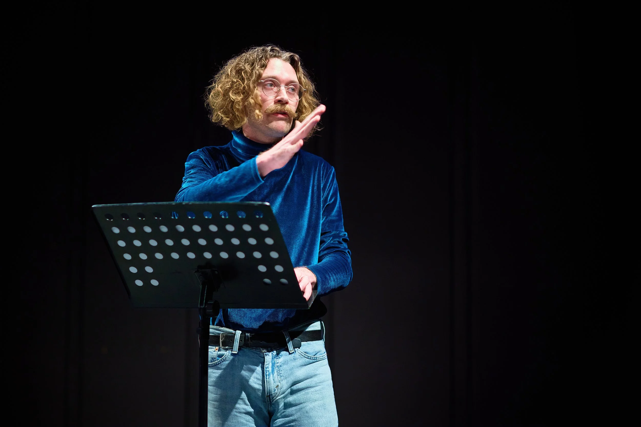 Un homme à moustache et cheveux bouclés, portant un pull en velours bleu, se tient devant un pupitre noir avec un trou perforé, faisant un geste avec sa main, sur fond noir.