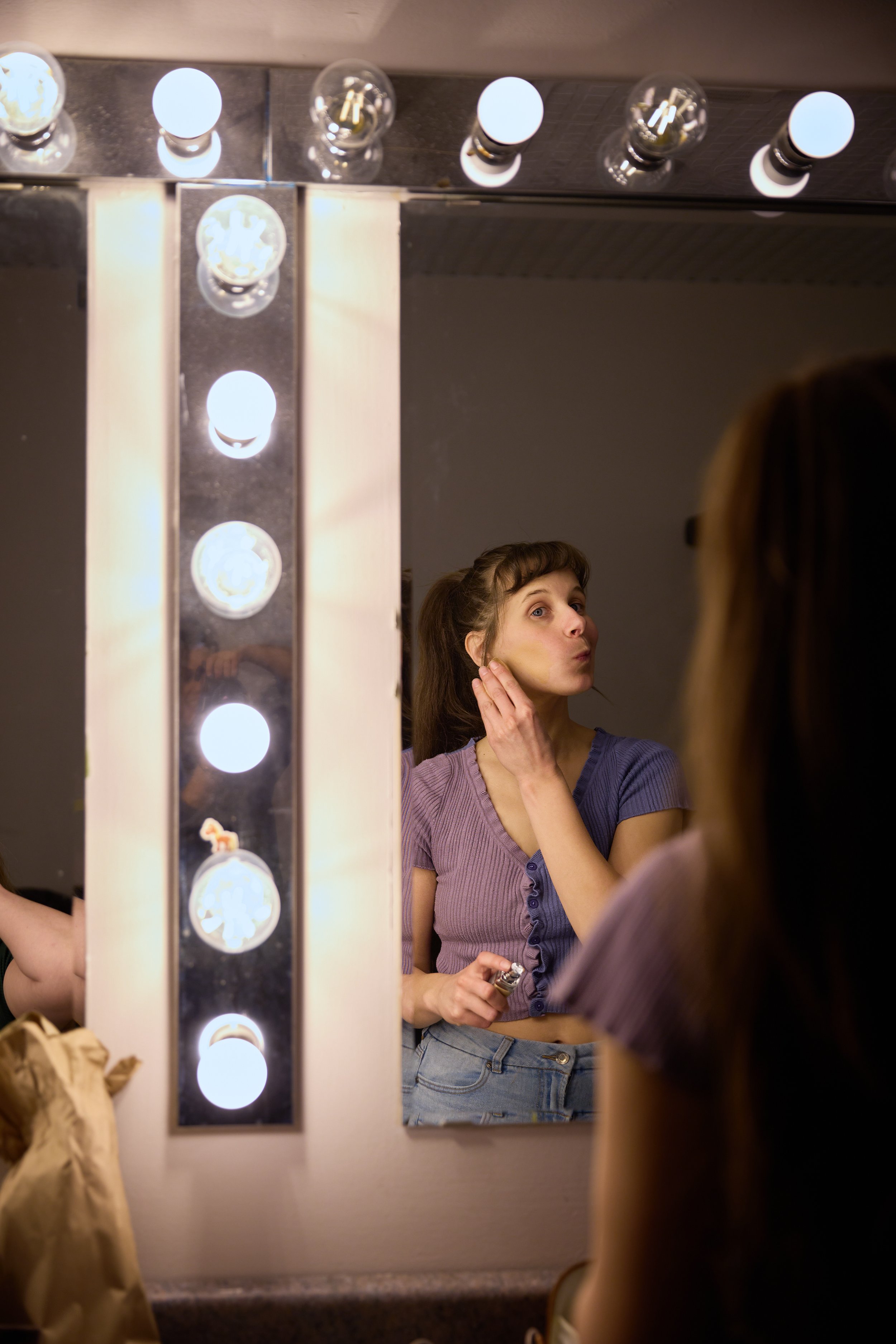 Une femme se regarde dans un miroir éclairé par des ampoules, en train de se maquiller ou de se préparer.