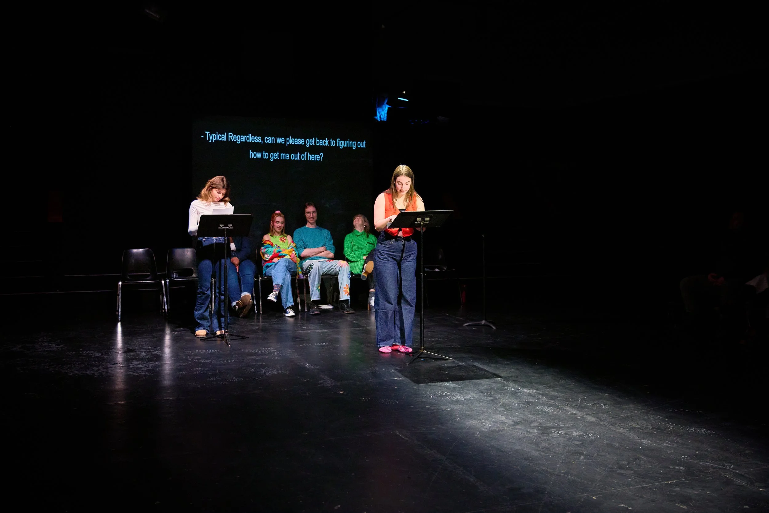 Une scène de théâtre avec six personnes sur scène, deux debout à des pupitres, quatre assises en arrière-plan, un écran noir en arrière-plan avec un texte en anglais, dans une salle sombre