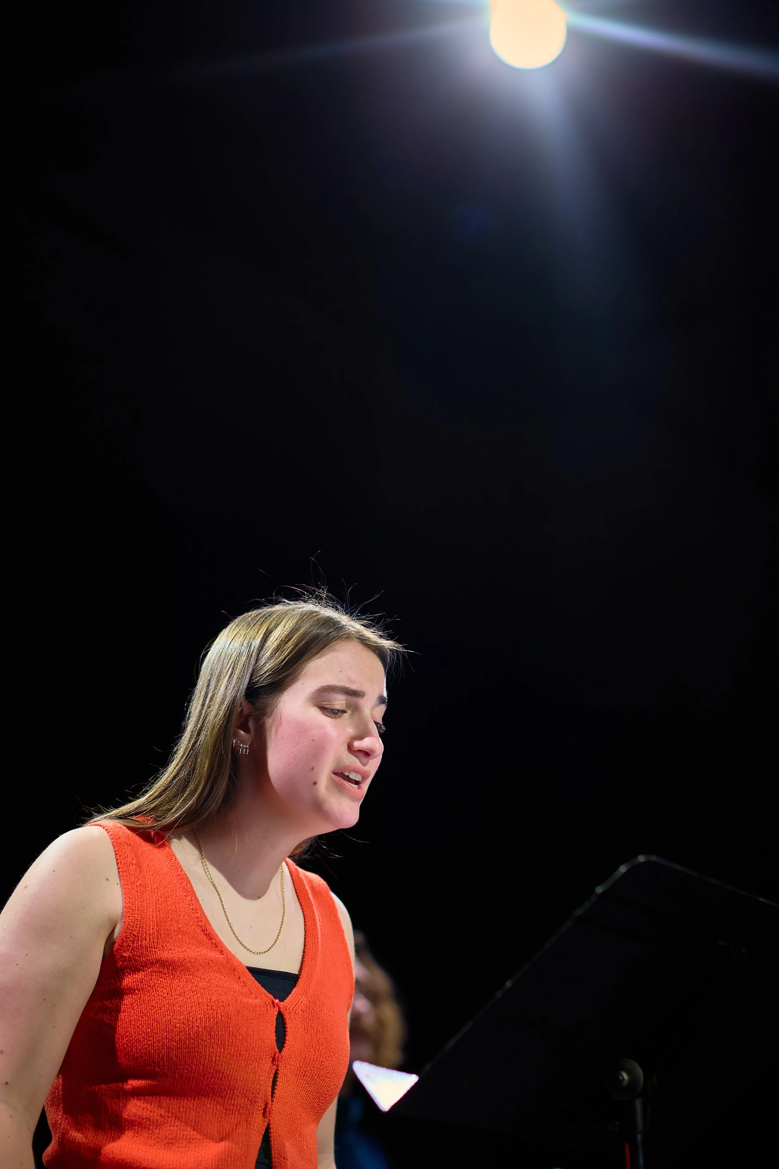 Une jeune femme chantant ou parlant, vêtue d'un top orange, se tient devant un pupitre ou microphone, sous un éclairage intense, avec un fond noir.