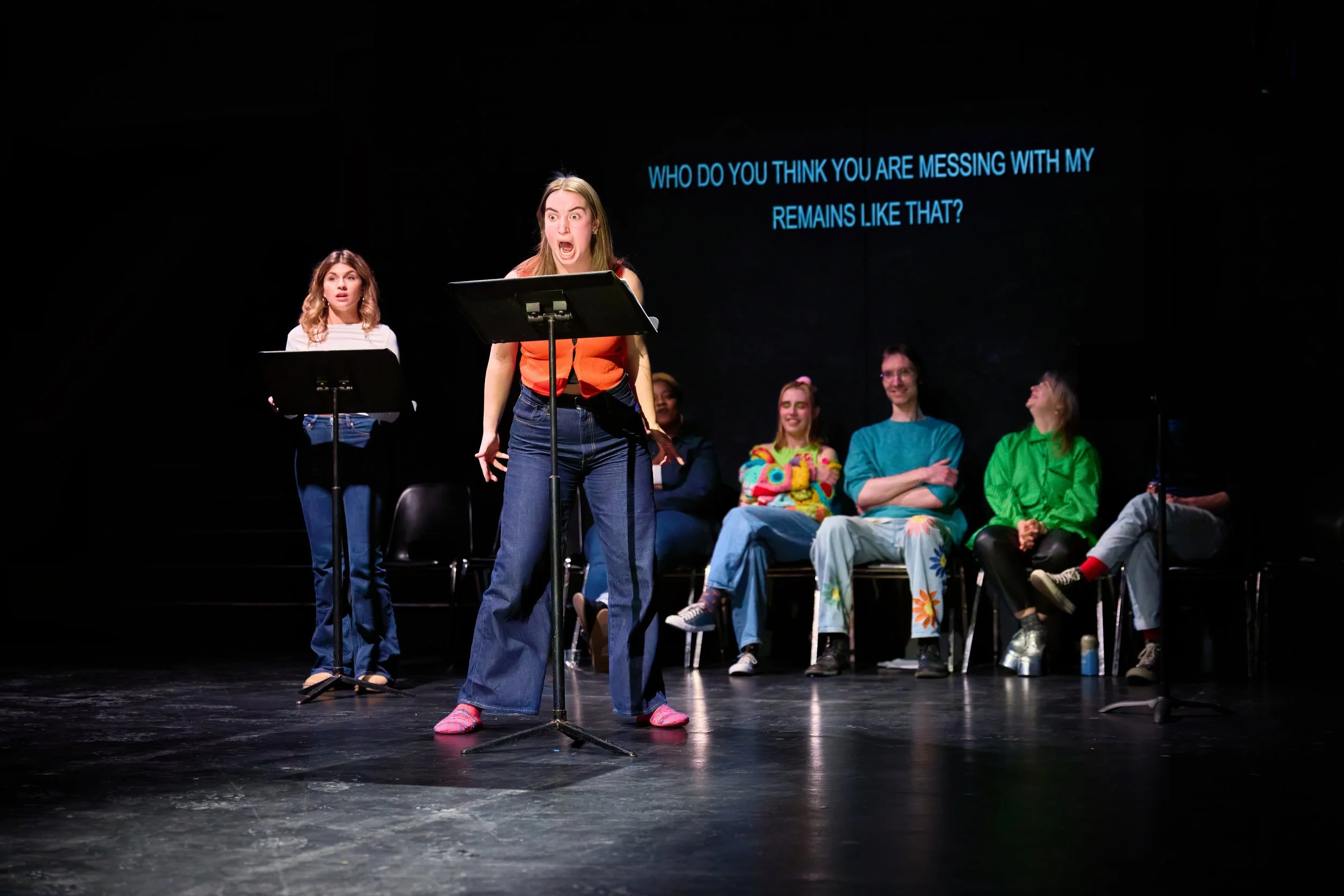 Une scène de théâtre avec plusieurs jeunes femmes, une au centre qui crie ou s'exprime avec passion, et une autre derrière qui regarde avec surprise. Des personnes sont assises sur des chaises en arrière-plan, et un texte en anglais apparaît à l'écra