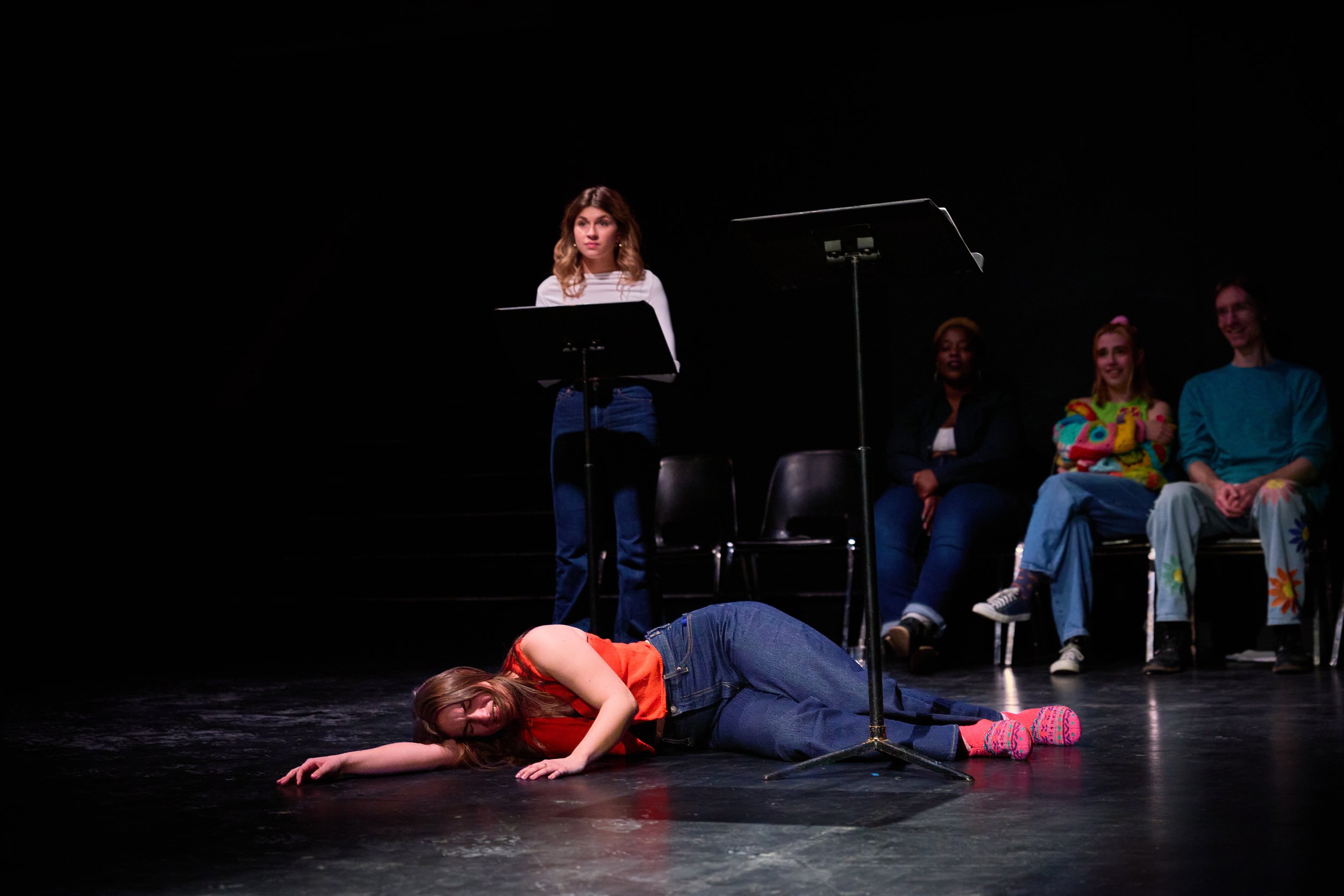 Une scène de théâtre avec une femme allongée sur le sol, une autre debout avec un pupitre, et trois personnes assises en arrière-plan, dont deux femmes et un homme, dans une ambiance sombre.