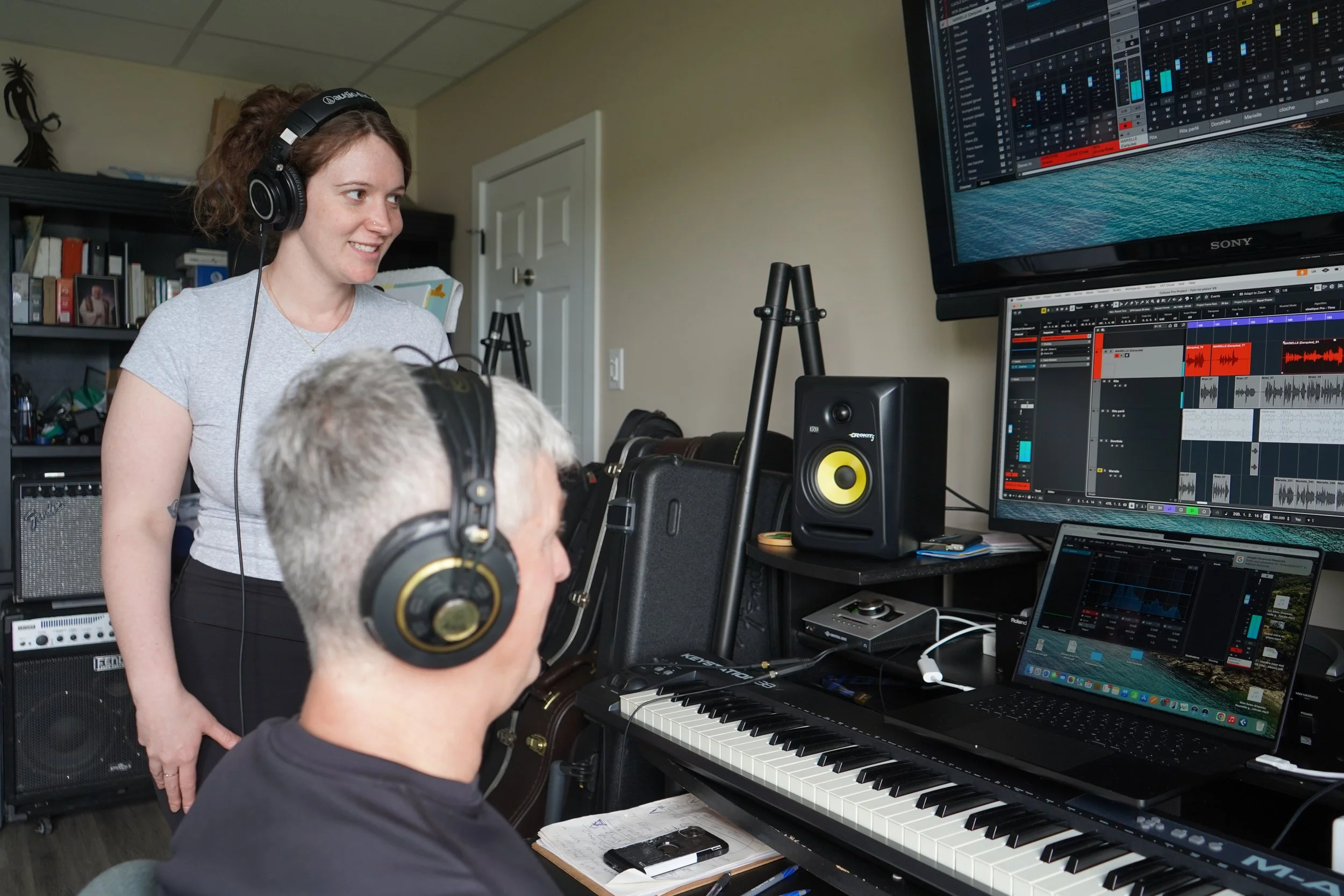 Une jeune femme avec un casque d'écoute sur la tête, regarde un homme âgé qui est en train de jouer du piano dans un studio d'enregistrement. Plusieurs écrans d'ordinateur affichent des logiciels de montage audio, et un clavier de piano est visible. 