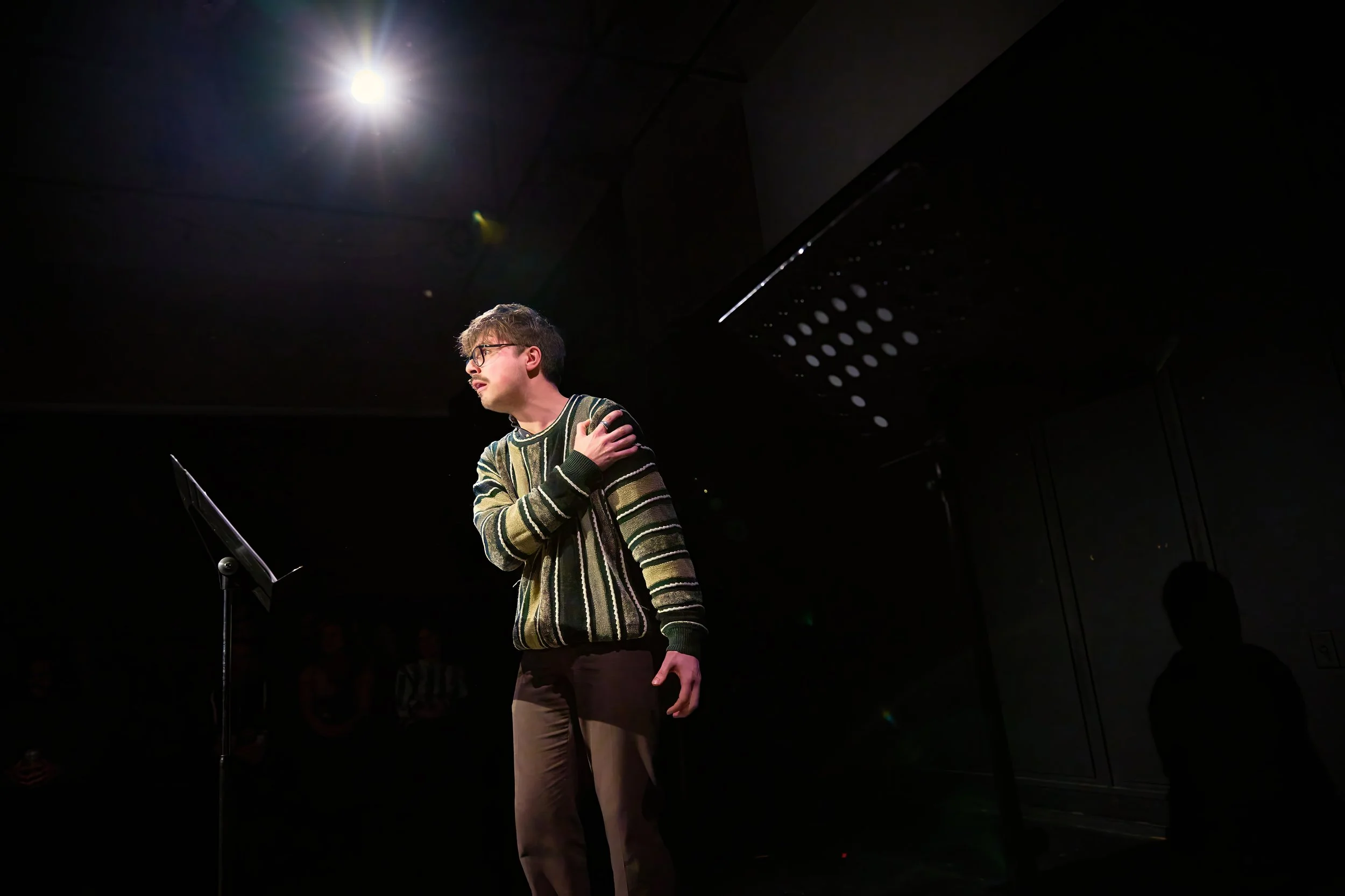 Un homme jeune, portant un pull rayé, en train de réciter ou de chanter sur scène, avec une main sur l'épaule, sous un éclairage dirigé vers lui dans une pièce sombre.