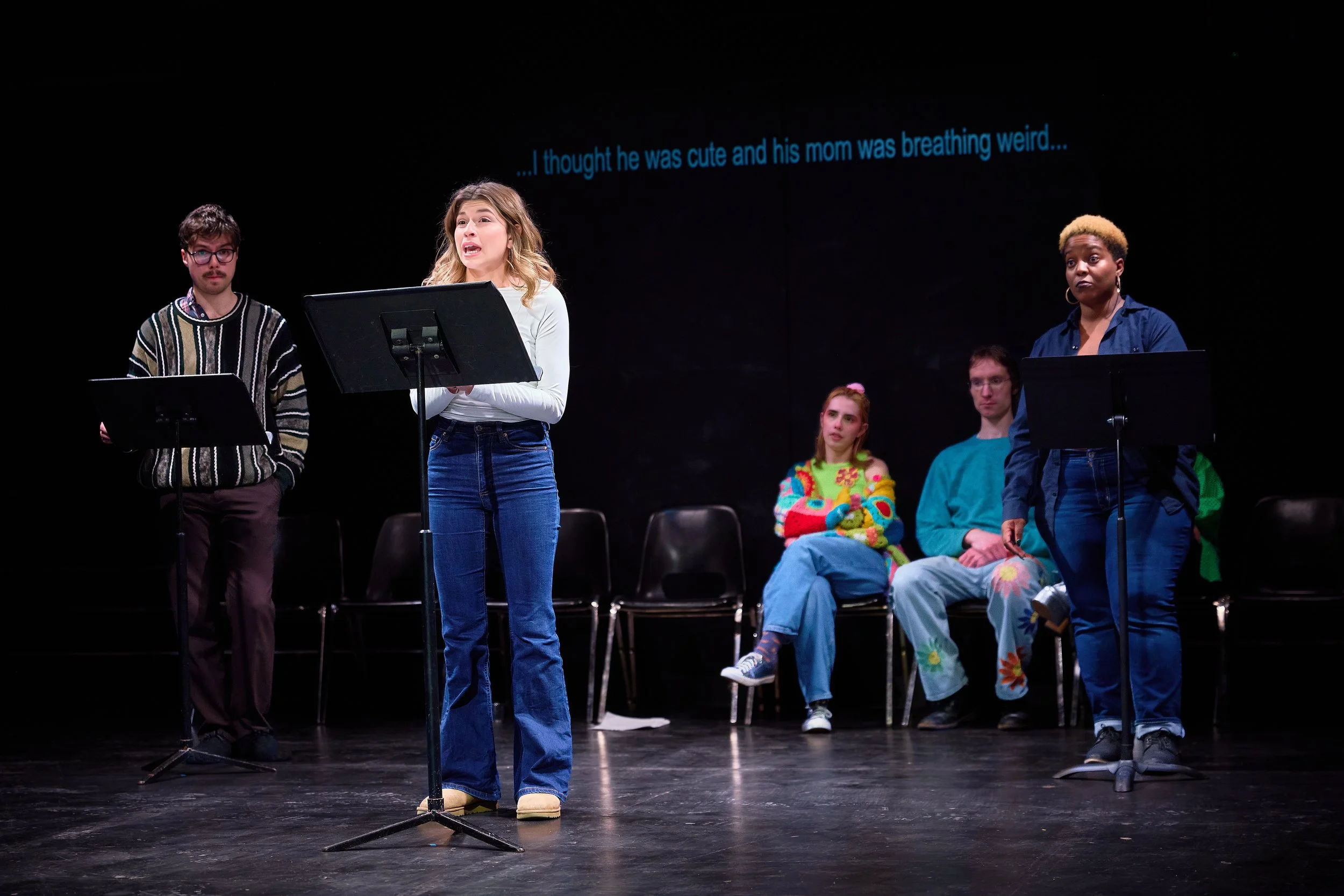 Groupe de jeunes acteurs en pleine scène de théâtre. Deux jeunes femmes à micro, en train de jouer, une avec une expression expressive. Quatre autres jeunes assis en arrière, regardant la scène, dont deux semblent intéressés. Fond noir avec texte en 