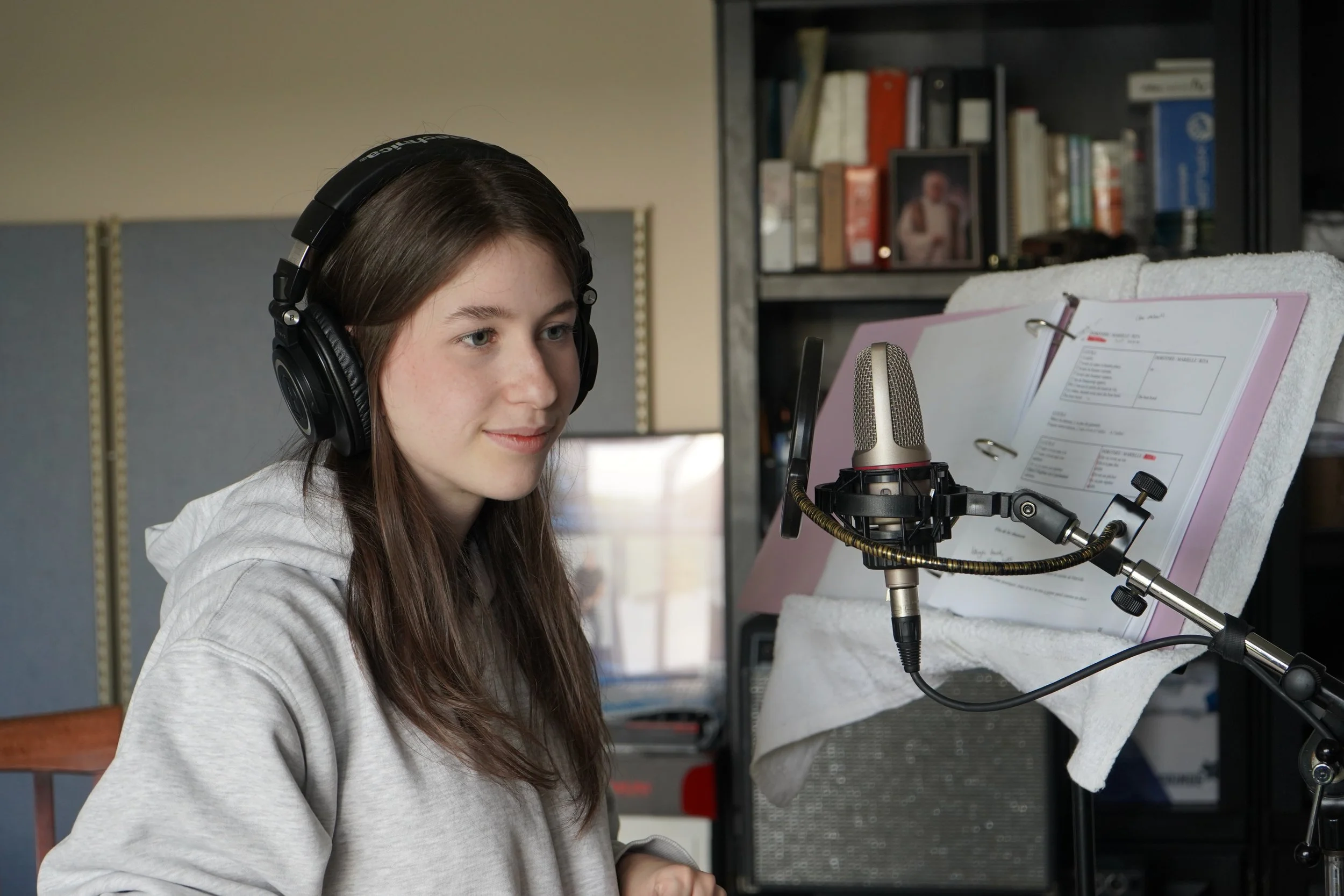 Une jeune fille portant un casque d'écoute travaille en studio d'enregistrement avec un micro et des partitions, dans une pièce avec des étagères contenant des livres en arrière-plan.
