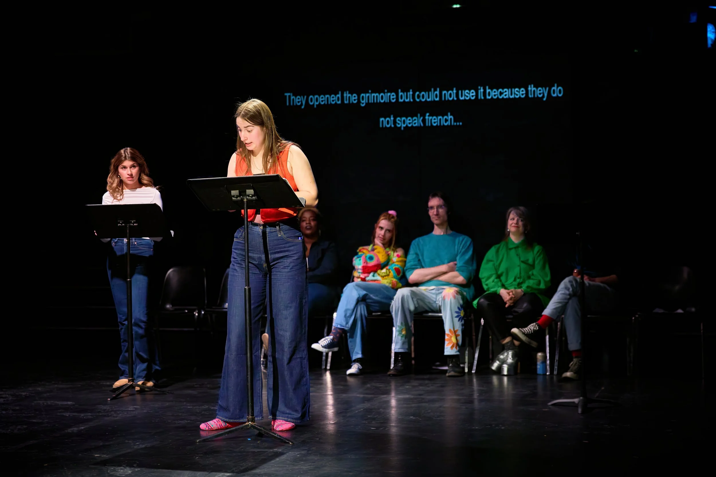 Une scène de théâtre avec plusieurs jeunes acteurs, une fille au centre lit un texte, tandis que les autres sont assis en arrière-plan. Un écran noir derrière affiche le texte en anglais: "They opened the grimoire but could not use it because they do