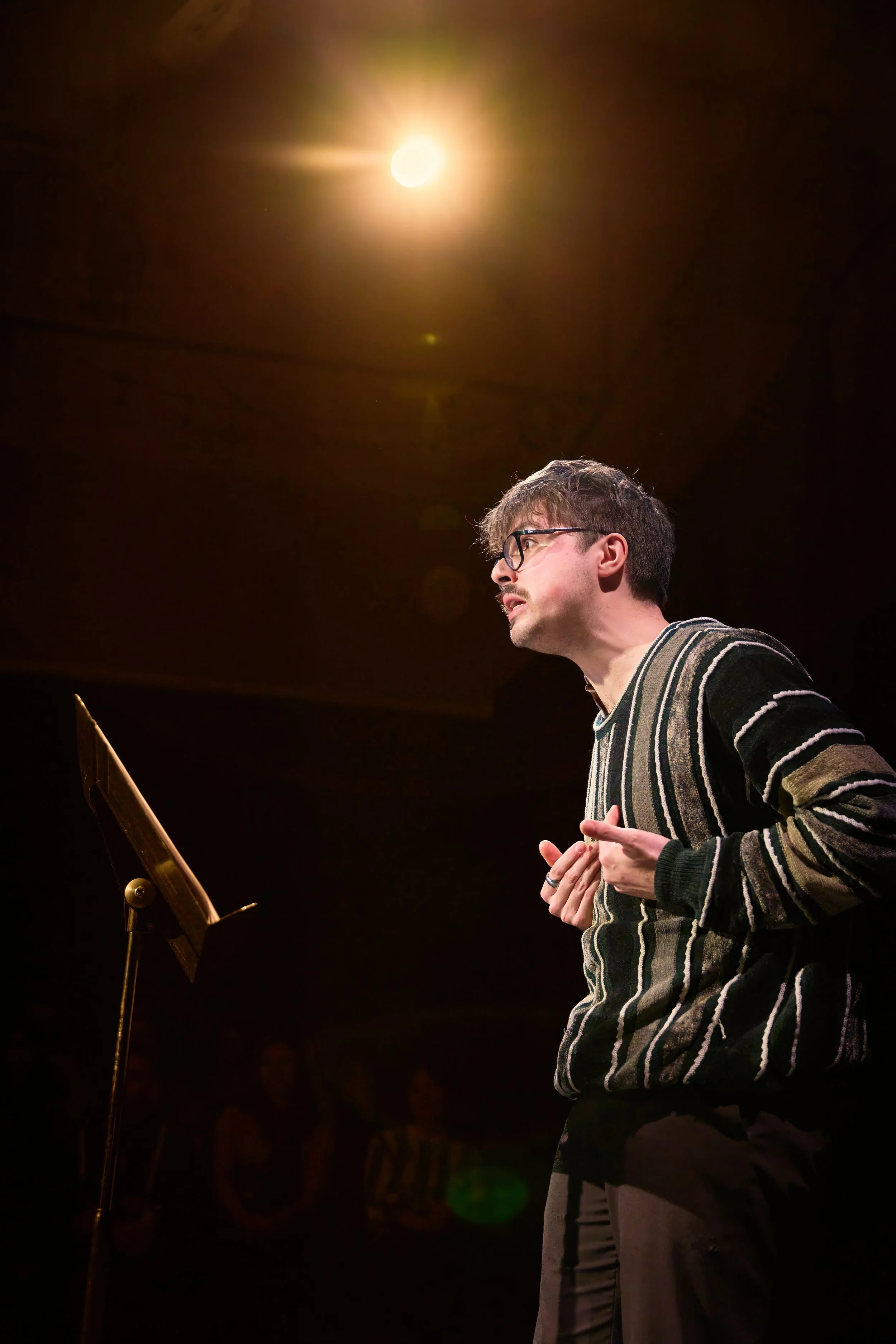 Un homme avec des lunettes, gesticulant et parlant devant un pupitre, sous un éclairage brillant dans une salle sombre.