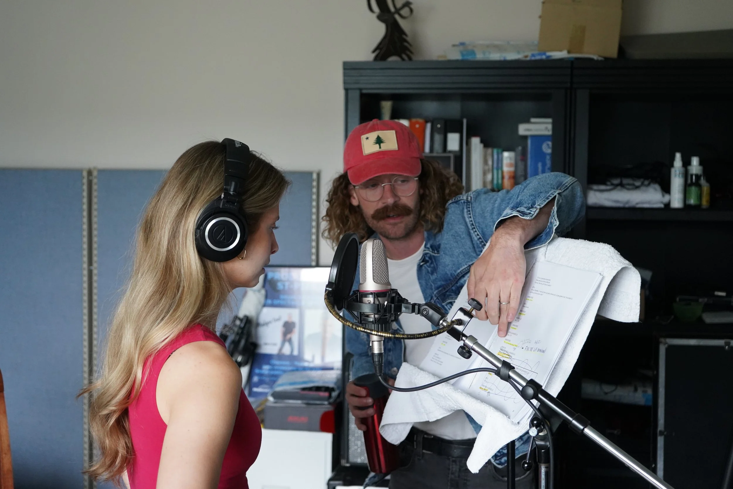 Deux personnes en studio d'enregistrement : une femme avec casque audio, une homme avec casquette rouge et lunettes, ajustant une feuille avec des notes et une ligne de micro.