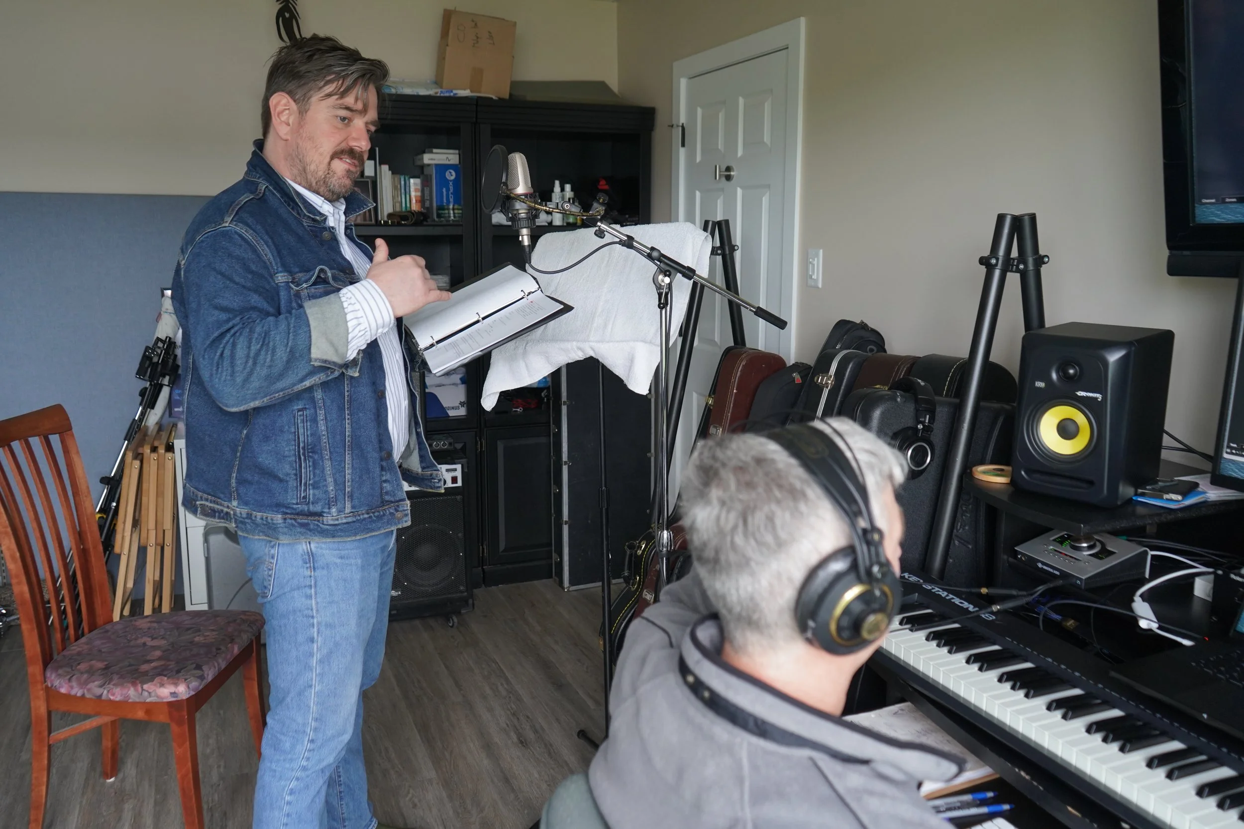 Un homme debout portant un carnet, un microphone et un support de caméra, semble enregistrer ou diriger une vidéo ou un podcast dans une pièce domestique, avec un autre homme assis jouant du piano.