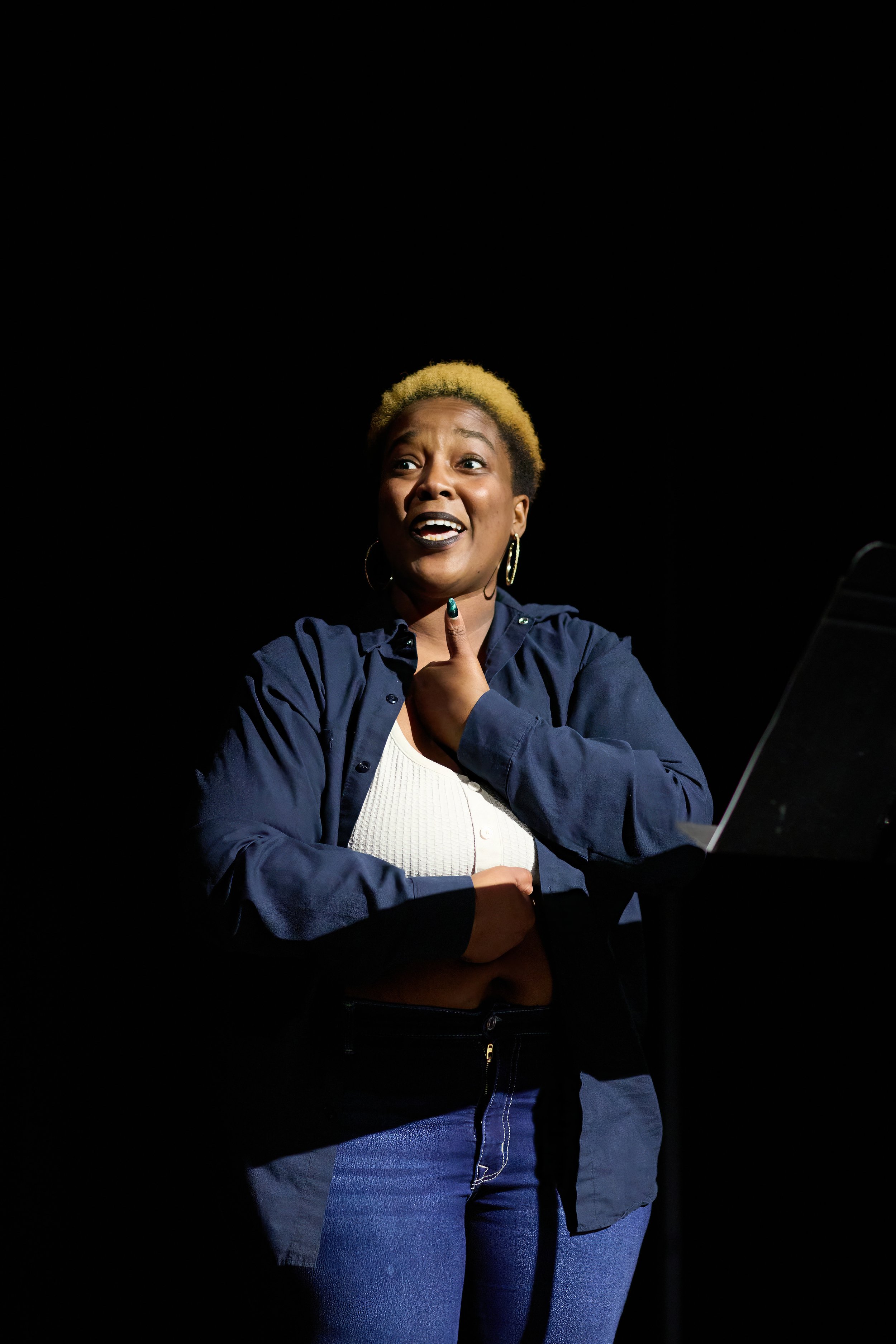 Une femme avec des cheveux blonds courts, portant une chemise bleue et une robe blanche, souriant et gesticulant lors d'une performance ou d'une présentation, avec un fond noir.