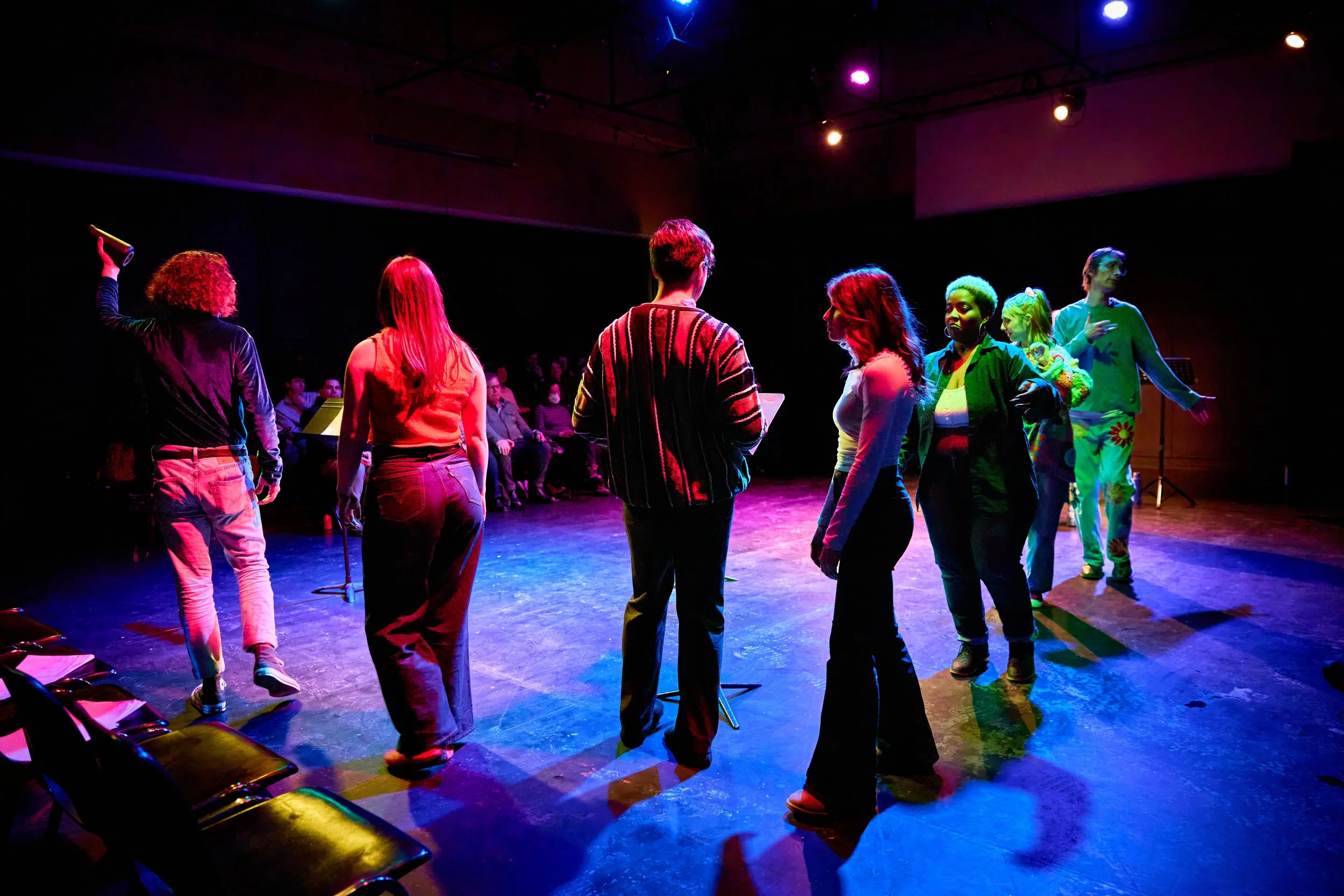 Un groupe de personnes, dans un espace sombre éclairé par des lumières colorées, semble préparer une performance ou une répétition, avec quelques spectateurs assis en arrière-plan.