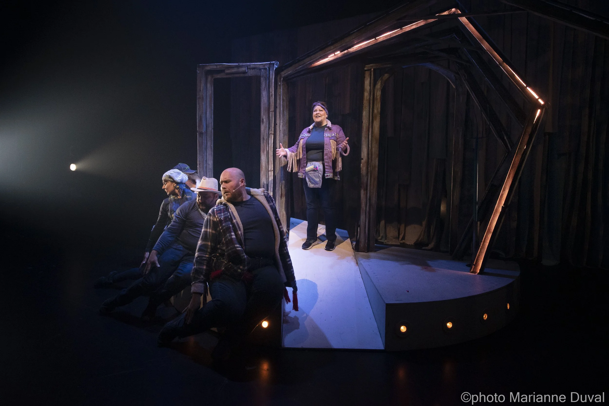 Des acteurs en costume sur scène, l'un d'eux parle, avec une structure en bois en arrière-plan.