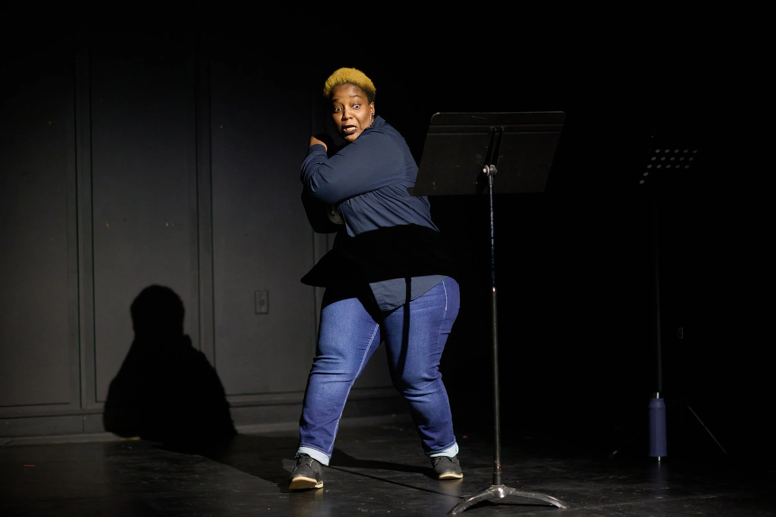 Une femme avec des cheveux blonds courts et une expression surprise se tenant sur scène derrière un pupitre de lecture, à côté d'un microphone, dans un environnement noir.
