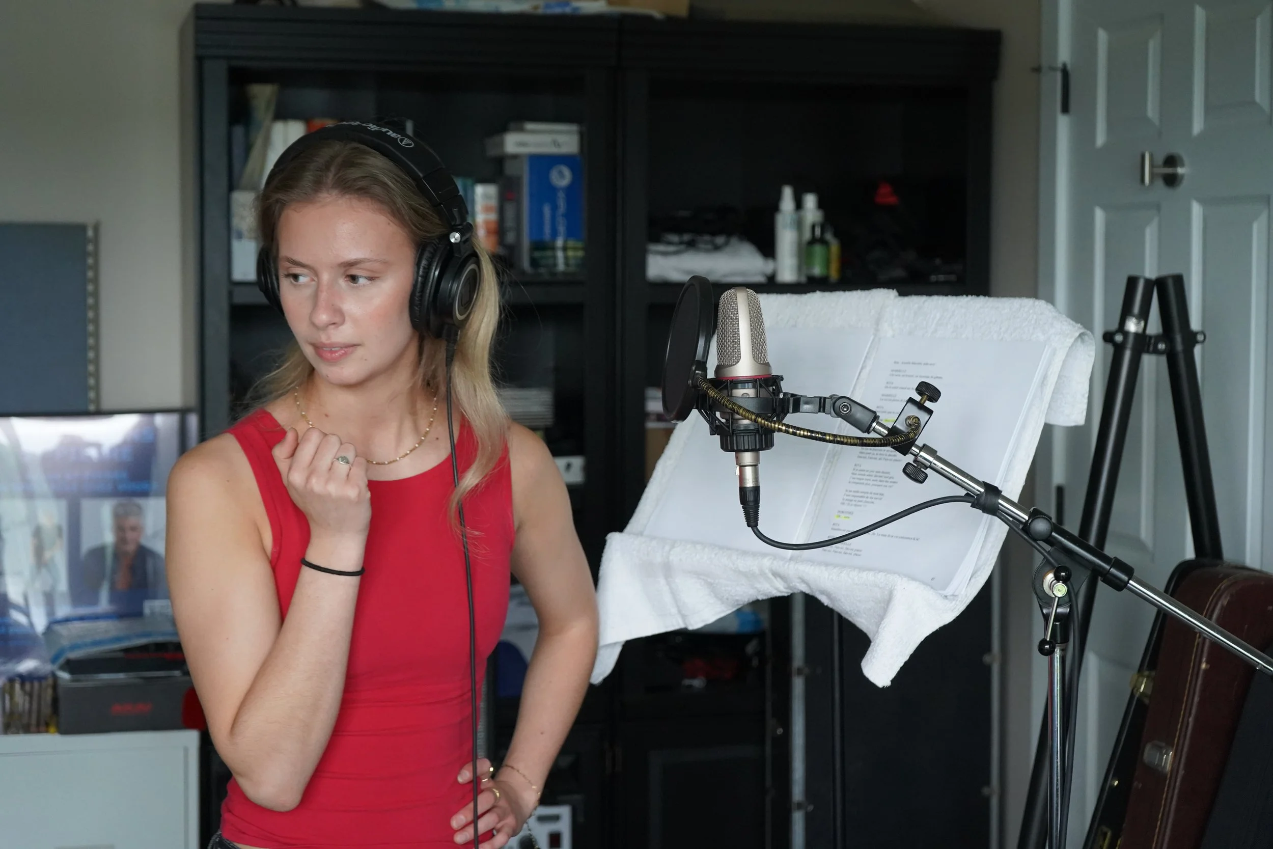 Une femme portant un t-shirt rouge et des écouteurs, enregistrement vocal avec un microphone en studio domestique.