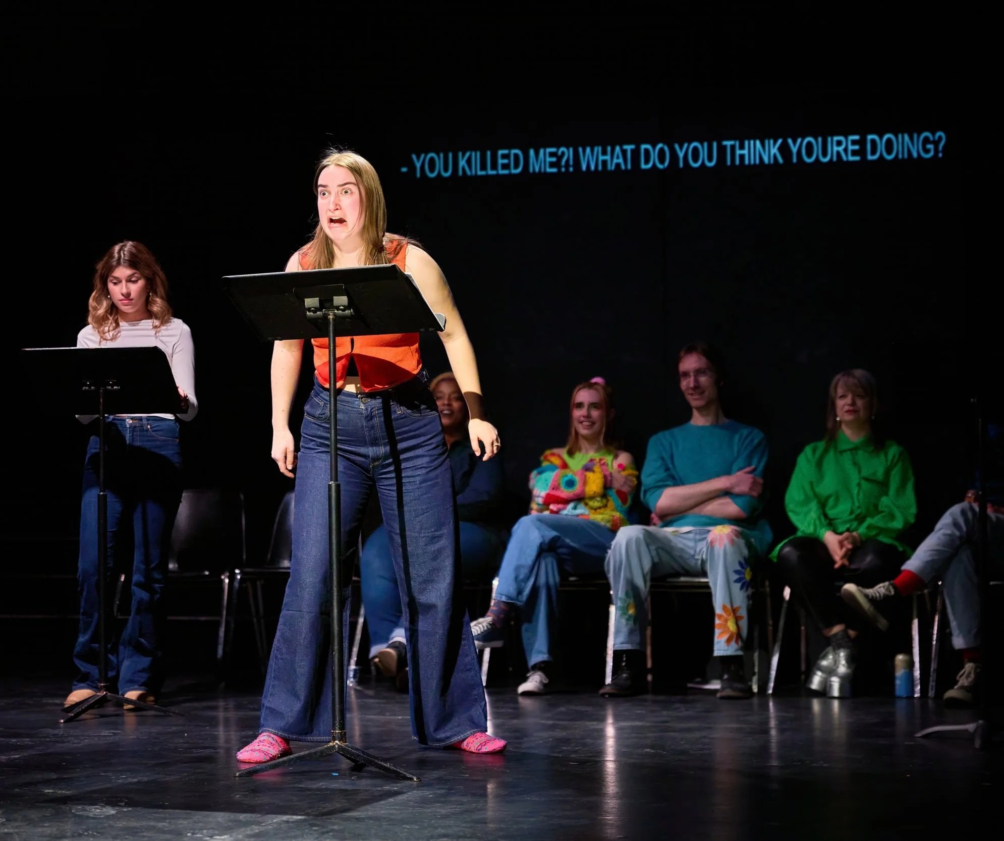 Une jeune fille debout sur scène, visiblement stressée ou inquiète, parlant dans un microphone, avec d'autres personnes assises en arrière-plan qui l'écoutent. Texte en arrière-plan en bleu : "- YOU KILLED ME?! WHAT DO YOU THINK YOU'RE DOING?".