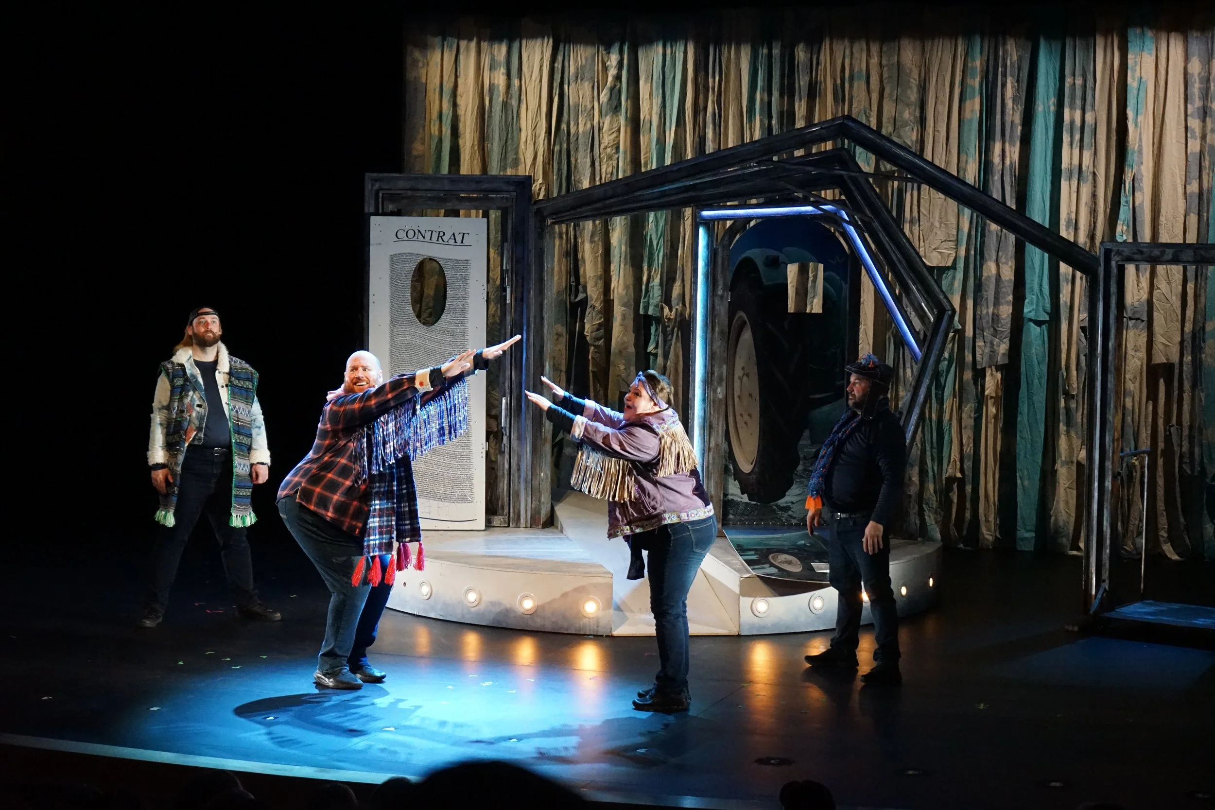 Scène de théâtre avec cinq acteurs en costumes dans une pièce avec un décor de forêt et une grande structure. Deux acteurs au centre se donnent la main, paraissant dans un échange dramatique, tandis que les autres les regardent. L’atmosphère est théâ