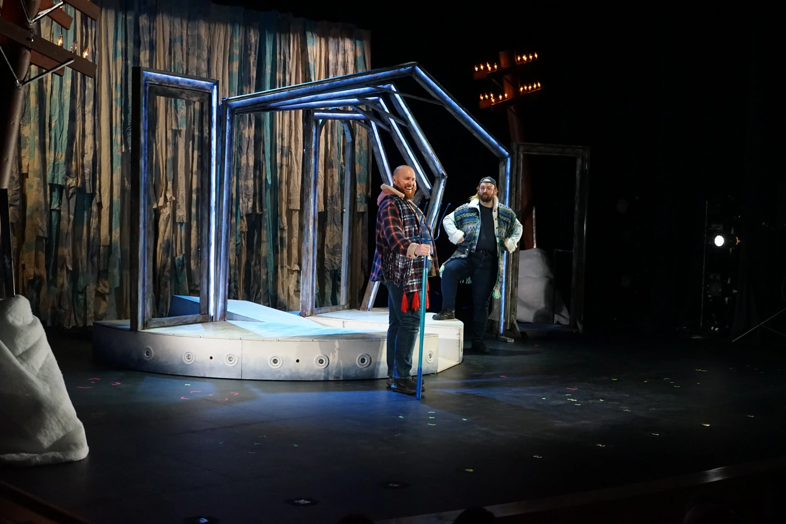 Deux hommes en costumes sur scène de théâtre avec un décor de cabane en bois et un mur de tissu en arrière-plan, l'un avec une canne et l'autre en position debout, en train de jouer une scène.